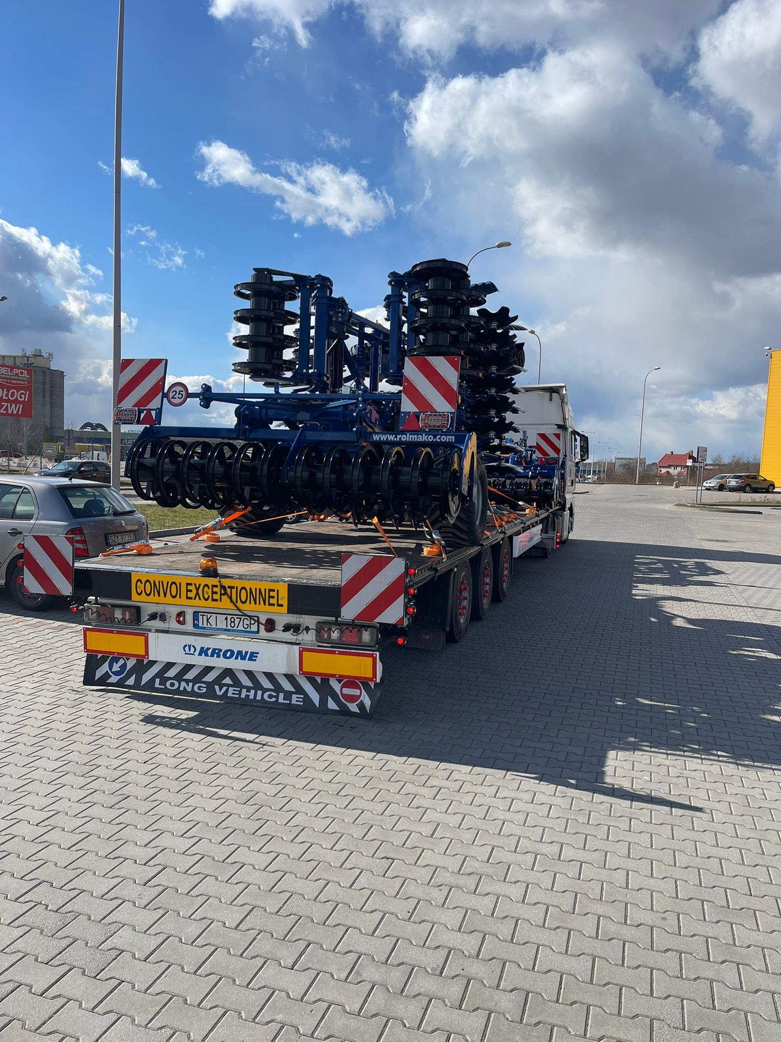 Transport niskopodwoziowy maszyny rolniczej (brona talerzowa) na naczepie z oznaczeniem 'CONVOI EXCEPTIONNEL' i ograniczeniem prędkości do 25 km/h.