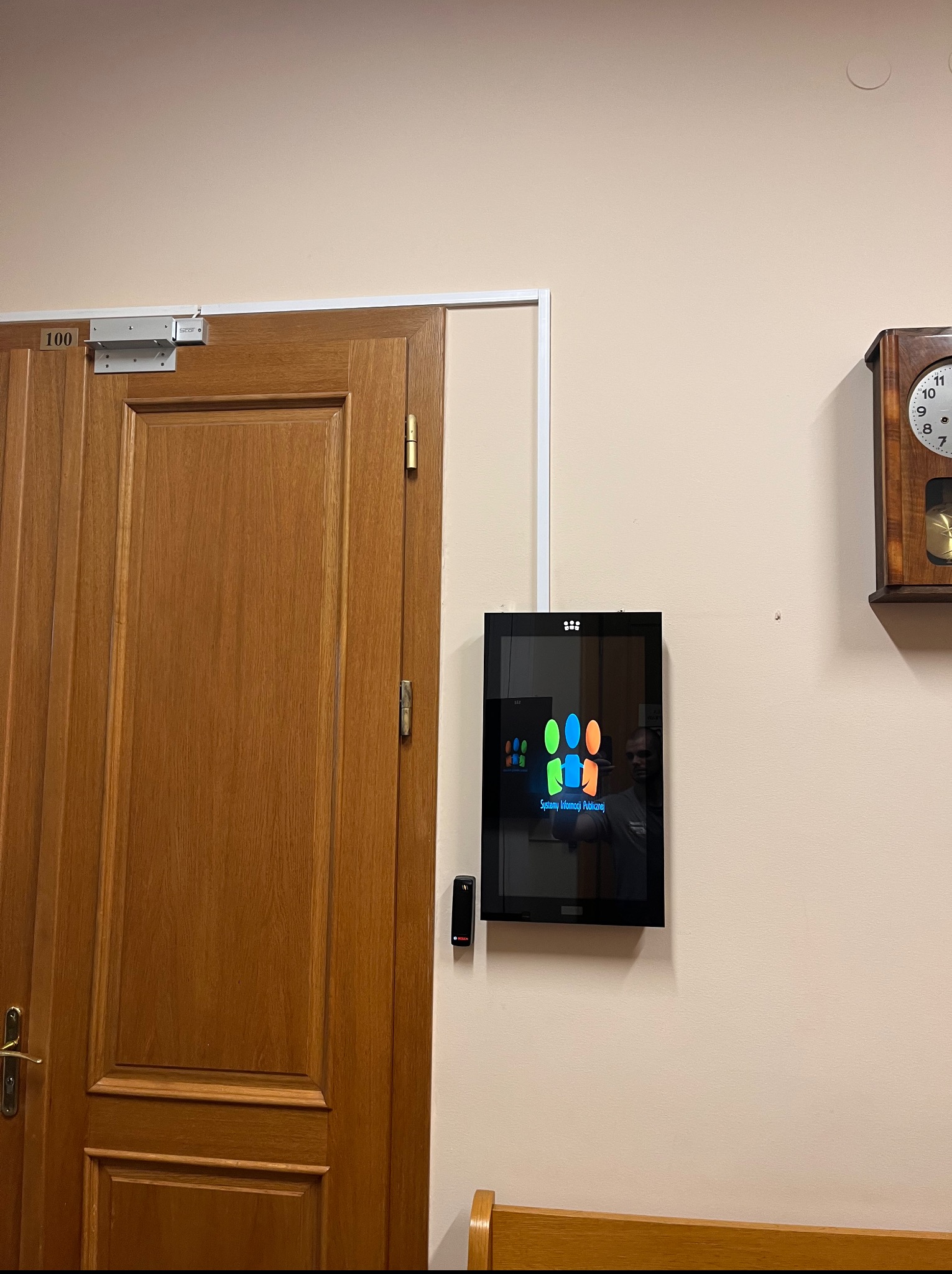 Panel informacyjny 'Systemy Informacji Publicznej' zamontowany na ścianie obok drewnianych drzwi z numerem 100 i zegarem. Widoczny czytnik kart dostępu.