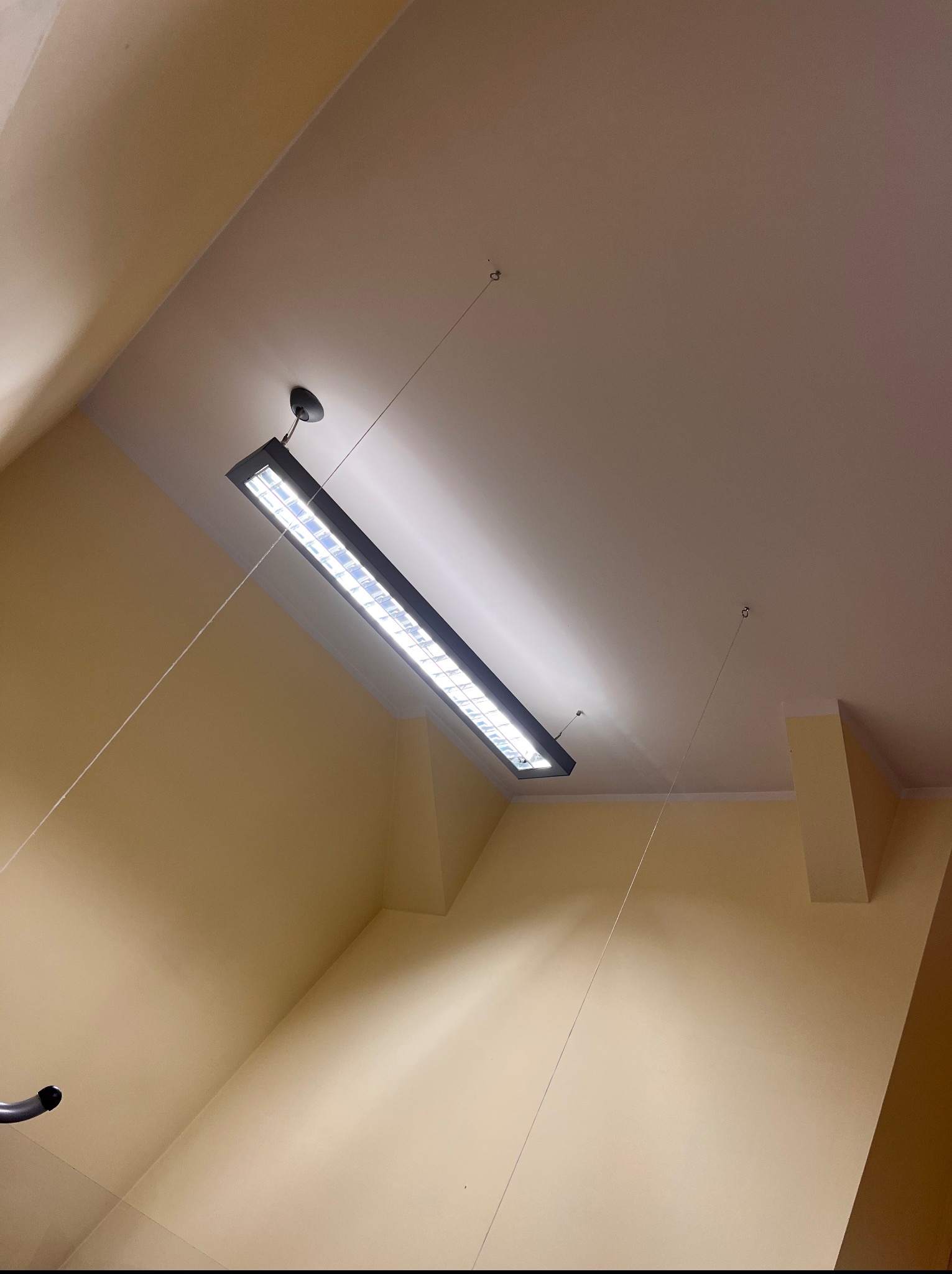 Zamontowana pod sufitem świetlówka w czarnej obudowie, zawieszona na linkach. Widok z dołu, żółte ściany w tle.