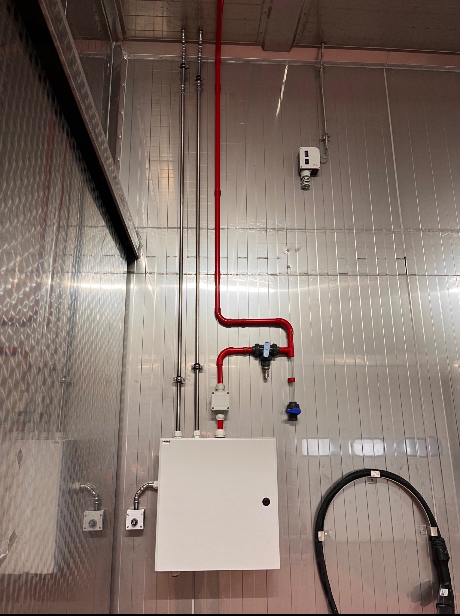 Metalowa ściana z instalacją: biała skrzynka, rury stalowe i czerwone rury z zaworami. Widoczne przewody i czujnik. Chłodnia lub pomieszczenie techniczne.
