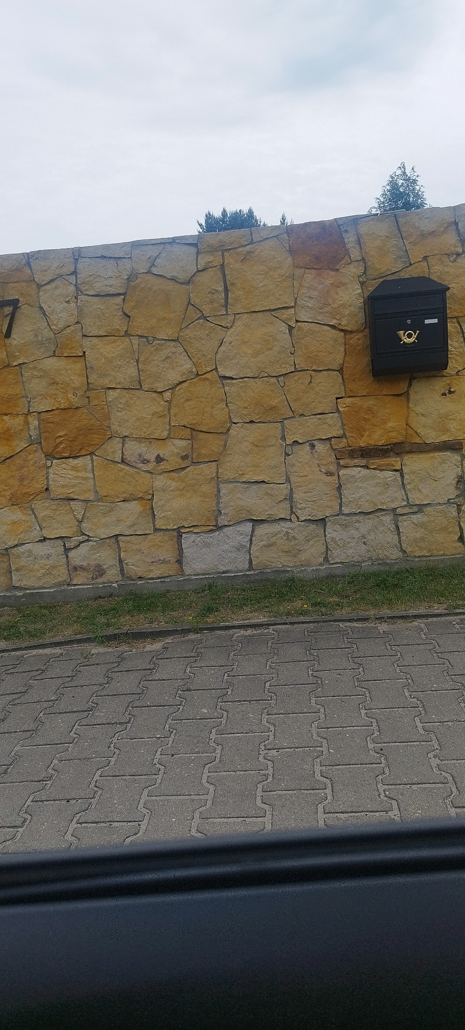 Kamienny mur z czarną skrzynką pocztową i numerem domu '7', widok z dołu na kostkę brukową i zieleń przy podstawie muru, pochmurne niebo w tle.