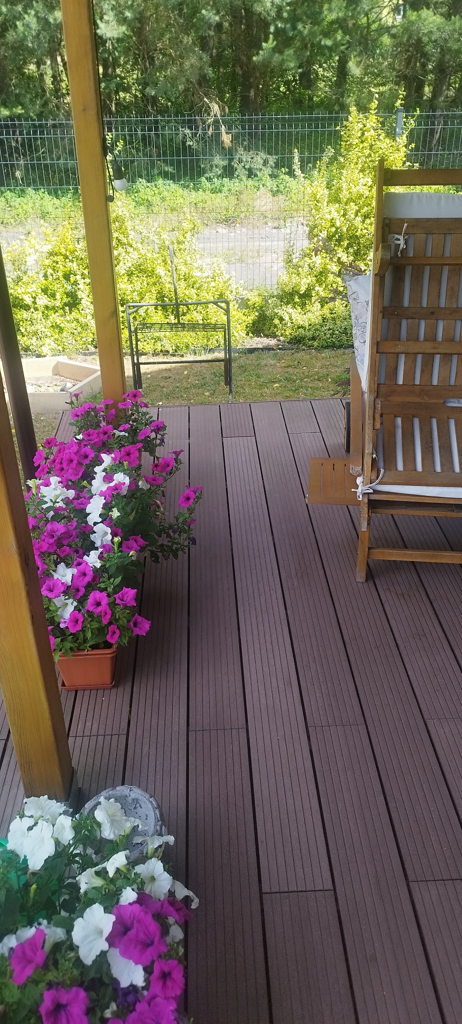 Kompozytowy taras w Warszawie z drewnianą leżanką i kwiatami w donicach, widok zadaszonego patio. Ciemne deski tarasowe tworzą przytulną przestrzeń.