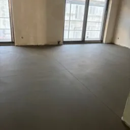 Szara, gładka posadzka w jasnym wnętrzu z dużymi oknami. Widoczne dylatacje posadzki. Ściany przygotowane do malowania. Realizacja w budynku wielorodzinnym.