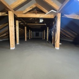 Posadzki Maszynowe Kujawa - Świeżo wylana, szara posadzka jastrychowa na poddaszu z drewnianą konstrukcją, widoczne słupy wspierające i kable zwisające z sufitu. Gotowa powierzchnia pod dalsze prace wykończeniowe.