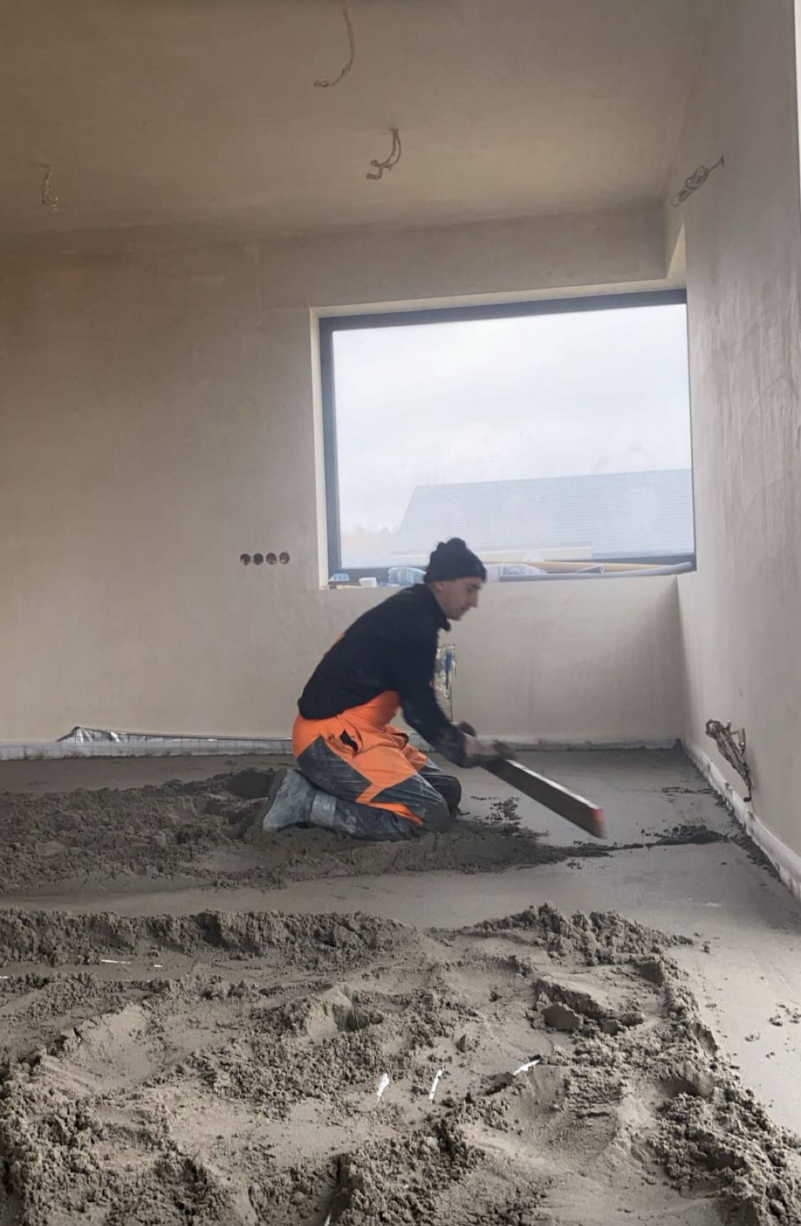 Pracownik w pomarańczowym kombinezonie wyrównuje szarą, mokrą masę na podłodze w jasnym pomieszczeniu z dużym oknem. Widoczne ślady po instalacjach na ścianach.