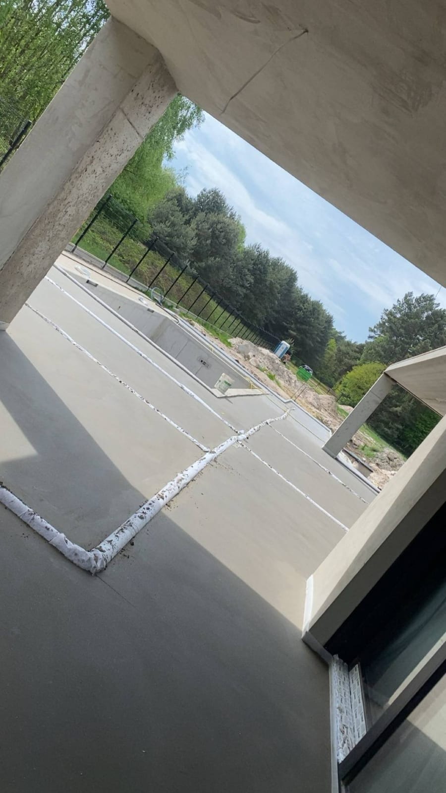 Świeżo wylana, gładka posadzka jastrychowa w nowym budynku, z widocznymi dylatacjami, otoczona zielenią w tle. Ujęcie z perspektywy narożnika pomieszczenia.