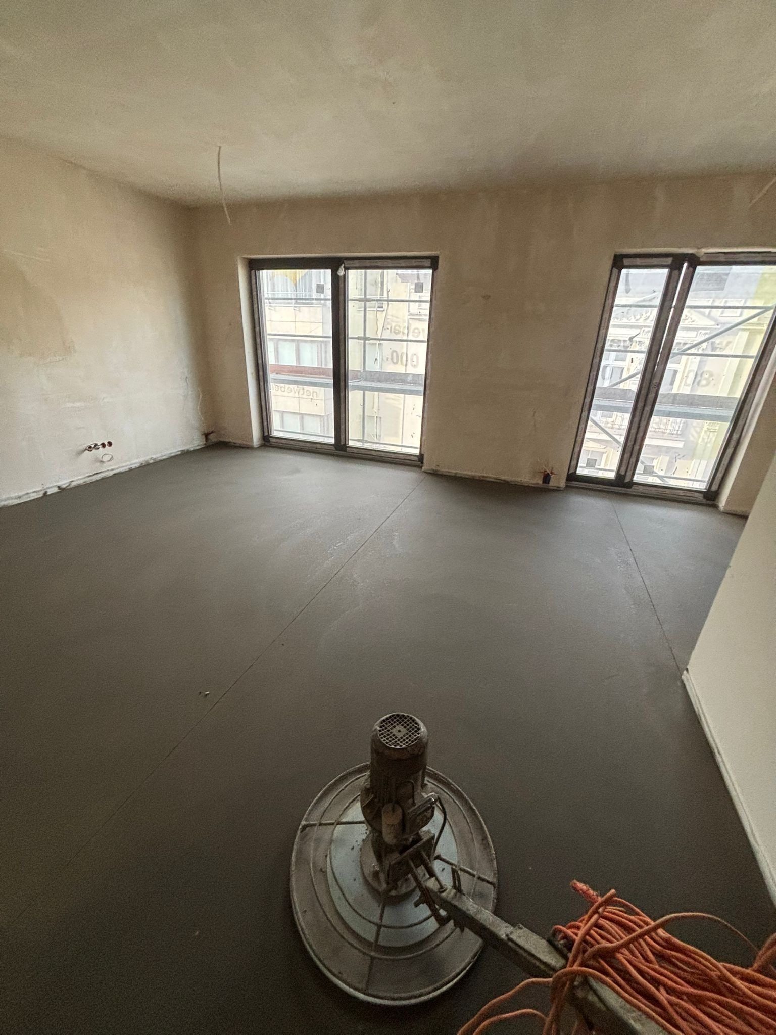 Wyrównywanie wylewki jastrychowej w jasnym pomieszczeniu z dwoma oknami, widoczna maszyna szlifierska i pomarańczowy kabel. Etap prac wykończeniowych.