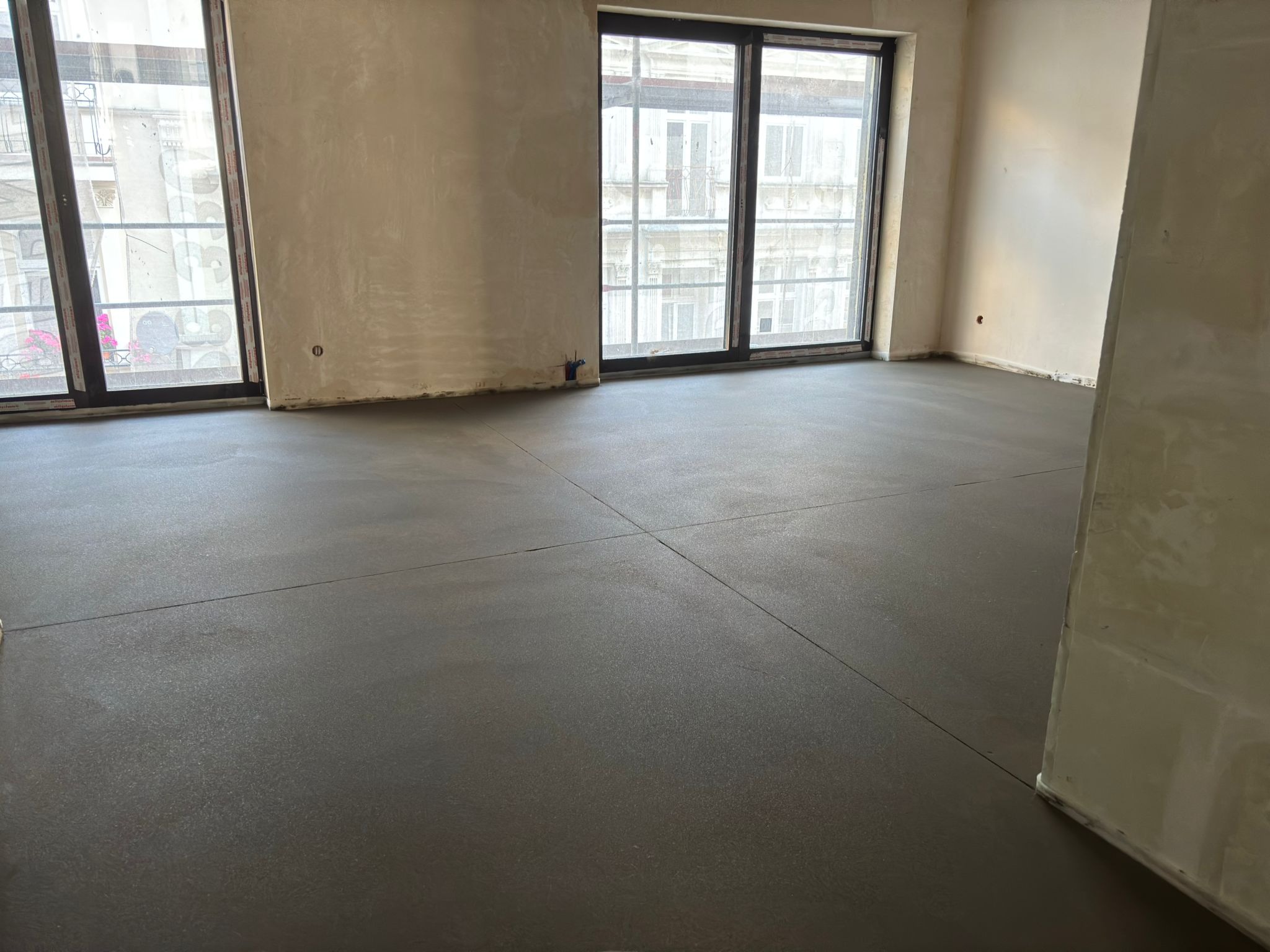 Gładka, ciemnoszara posadzka jastrychowa w jasnym wnętrzu z dużymi oknami balkonowymi. Widoczne dylatacje. Ściany w stanie surowym, przygotowane do dalszych prac.