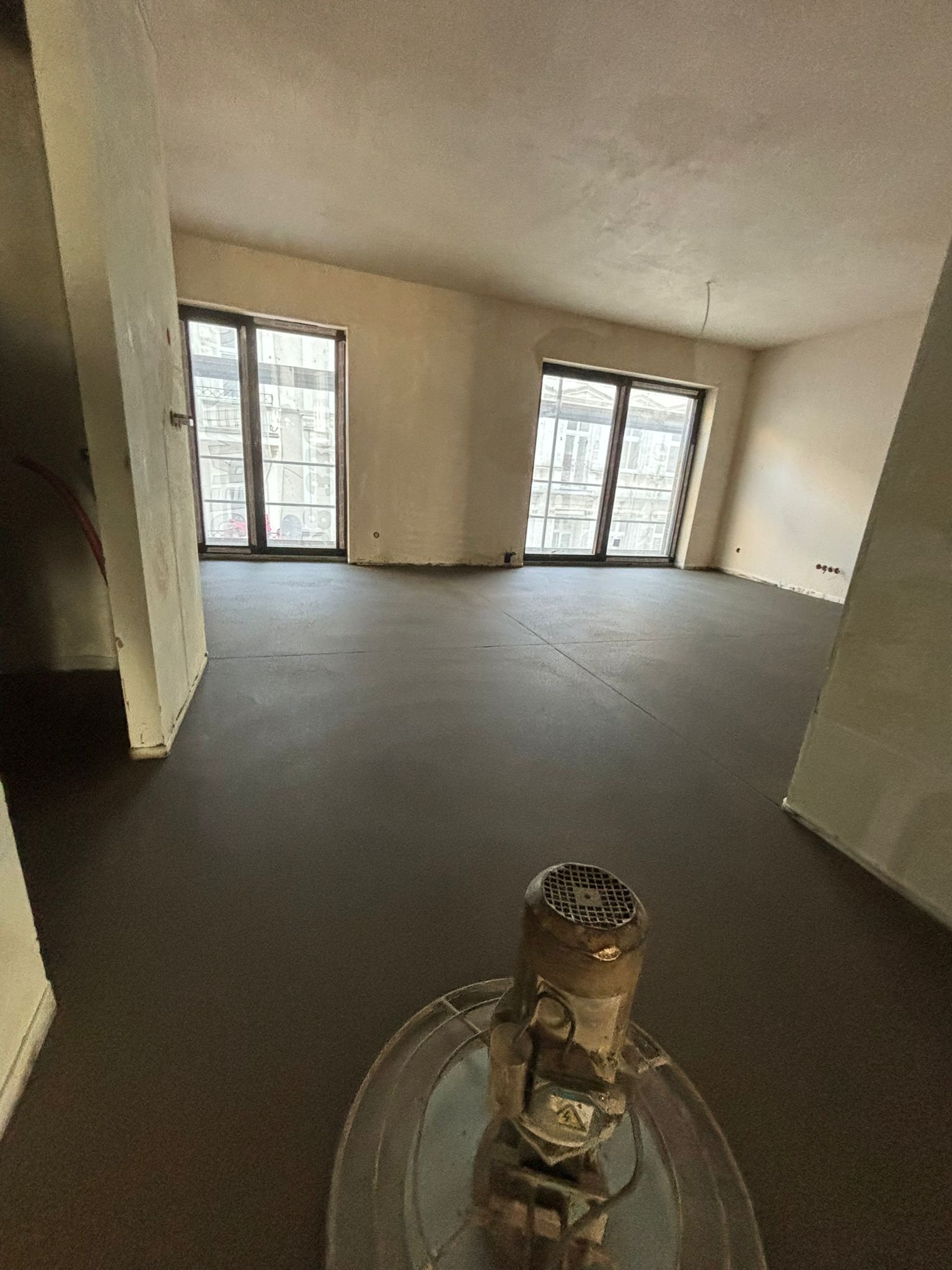 Wyrównana, ciemnoszara posadzka jastrychowa w jasnym wnętrzu z dwoma oknami. W tle widoczna maszyna do wygładzania powierzchni.