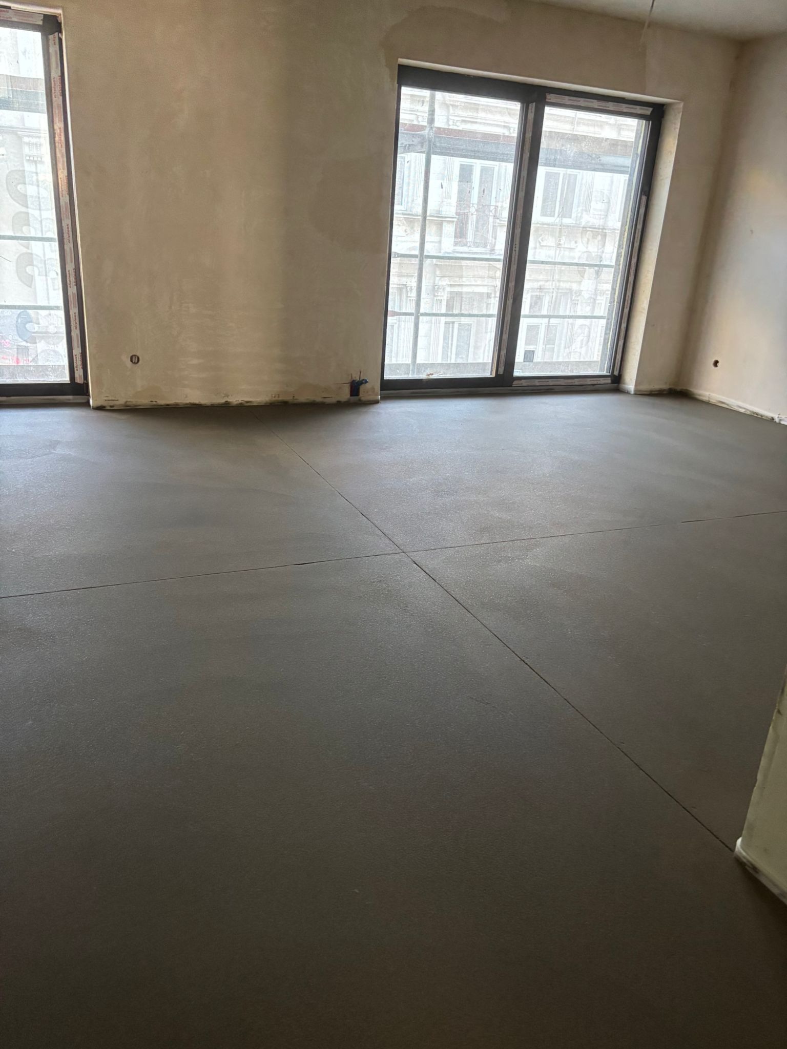 Szara, gładka posadzka w jasnym wnętrzu z dużymi oknami. Widoczne dylatacje posadzki. Ściany przygotowane do malowania. Realizacja w budynku wielorodzinnym.