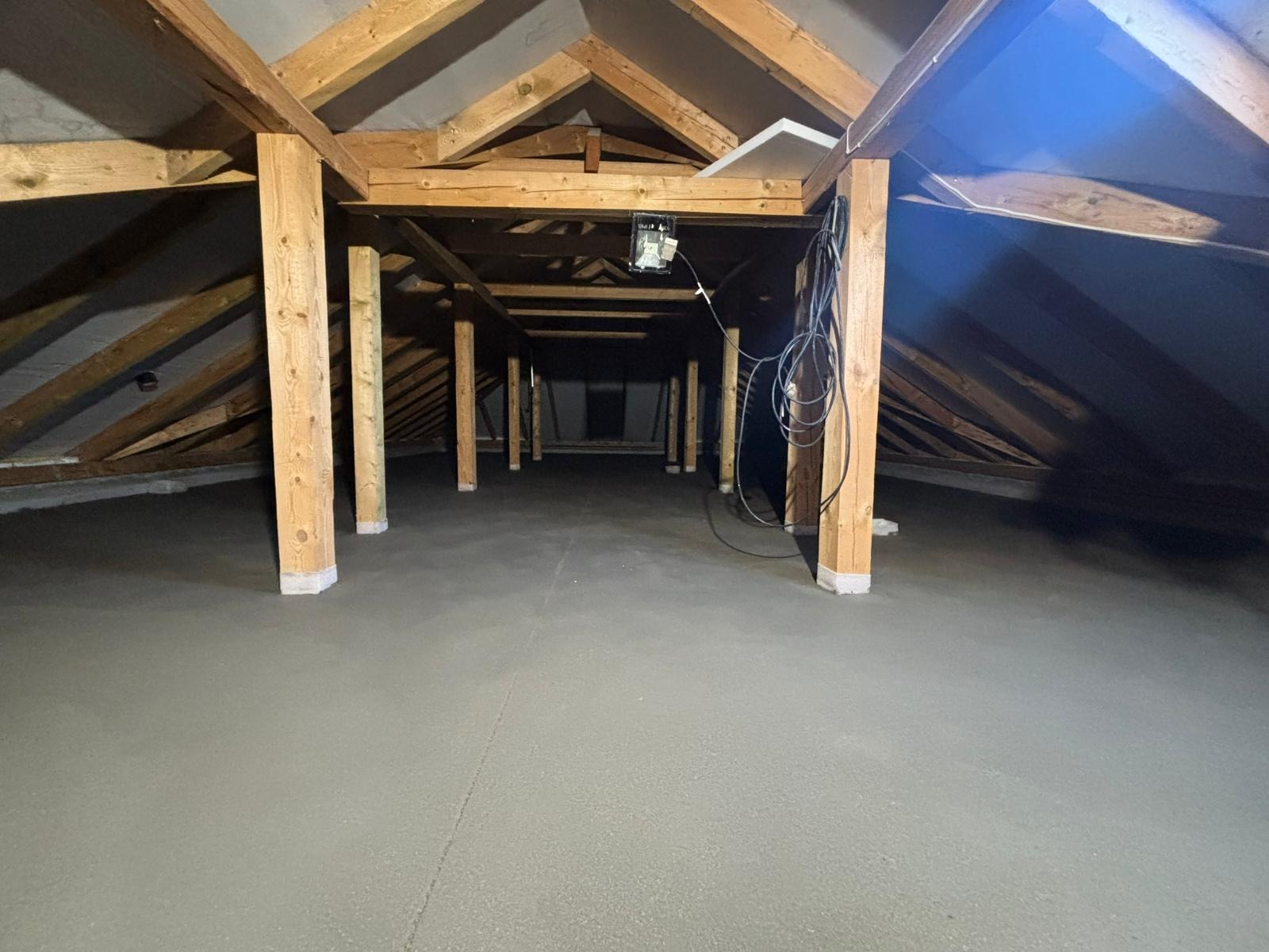 Świeżo wylana, szara posadzka jastrychowa na poddaszu z drewnianą konstrukcją, widoczne słupy wspierające i kable zwisające z sufitu. Gotowa powierzchnia pod dalsze prace wykończeniowe.