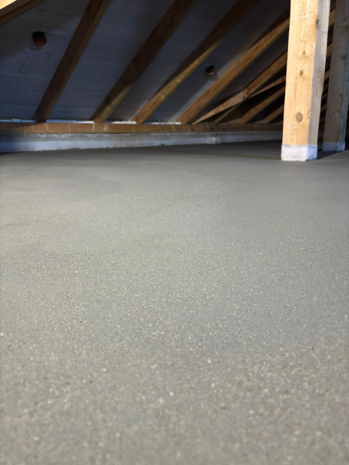 Szara, świeżo wylana posadzka jastrychowa w poddaszu, z widocznymi drewnianymi elementami konstrukcyjnymi dachu i słupem wspierającym.