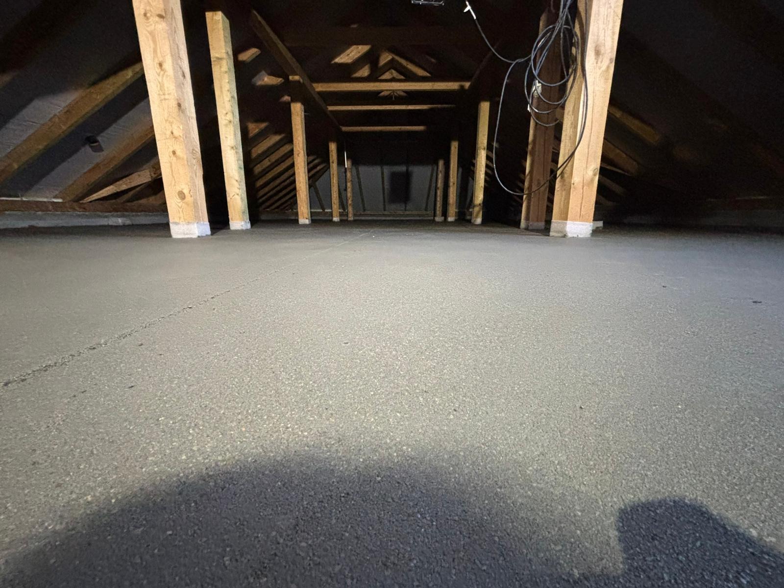 Szary jastrych w surowym wnętrzu poddasza z drewnianą konstrukcją i słupami. Widok z poziomu podłogi w głąb pomieszczenia.