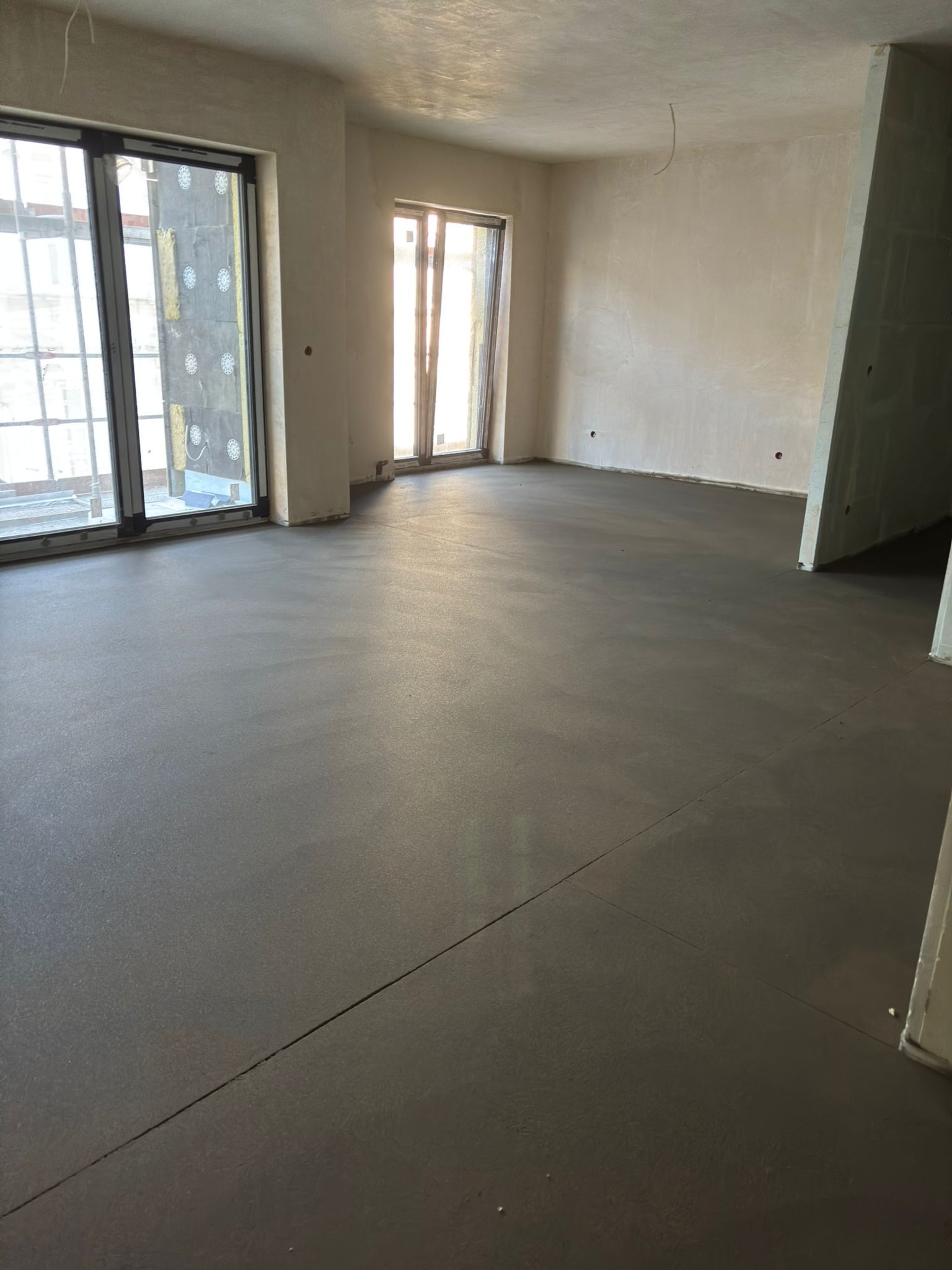 Wylana, gładka posadzka jastrychowa w jasnym pomieszczeniu z dużymi oknami. Widoczne dylatacje. Świeżo wykonana wylewka podłogowa.
