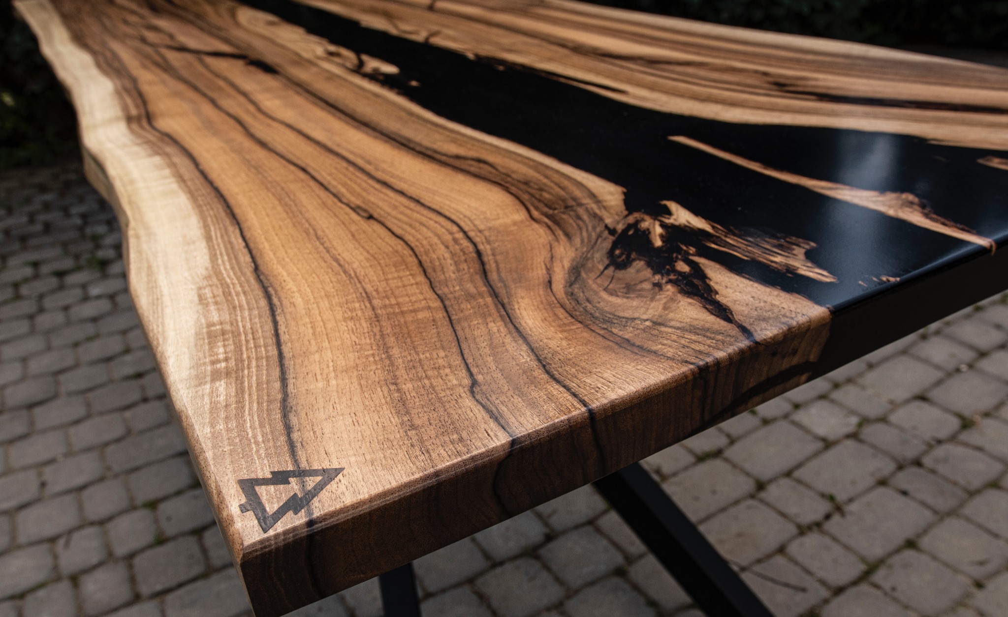 Blat stołu z żywicą epoksydową, ukazujący unikalne usłojenie drewna i czarną żywicę. Nierówna krawędź, logo w rogu. Metalowa podstawa, kostka brukowa w tle.