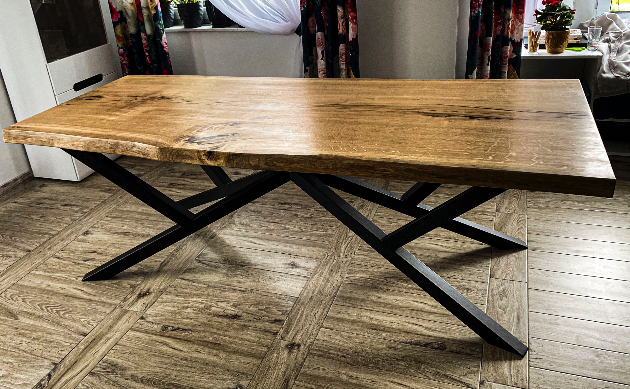 Stół z blatem z litego drewna o nieregularnych krawędziach, wsparty na czarnej, geometrycznej podstawie. Parkiet w odcieniach brązu w tle.