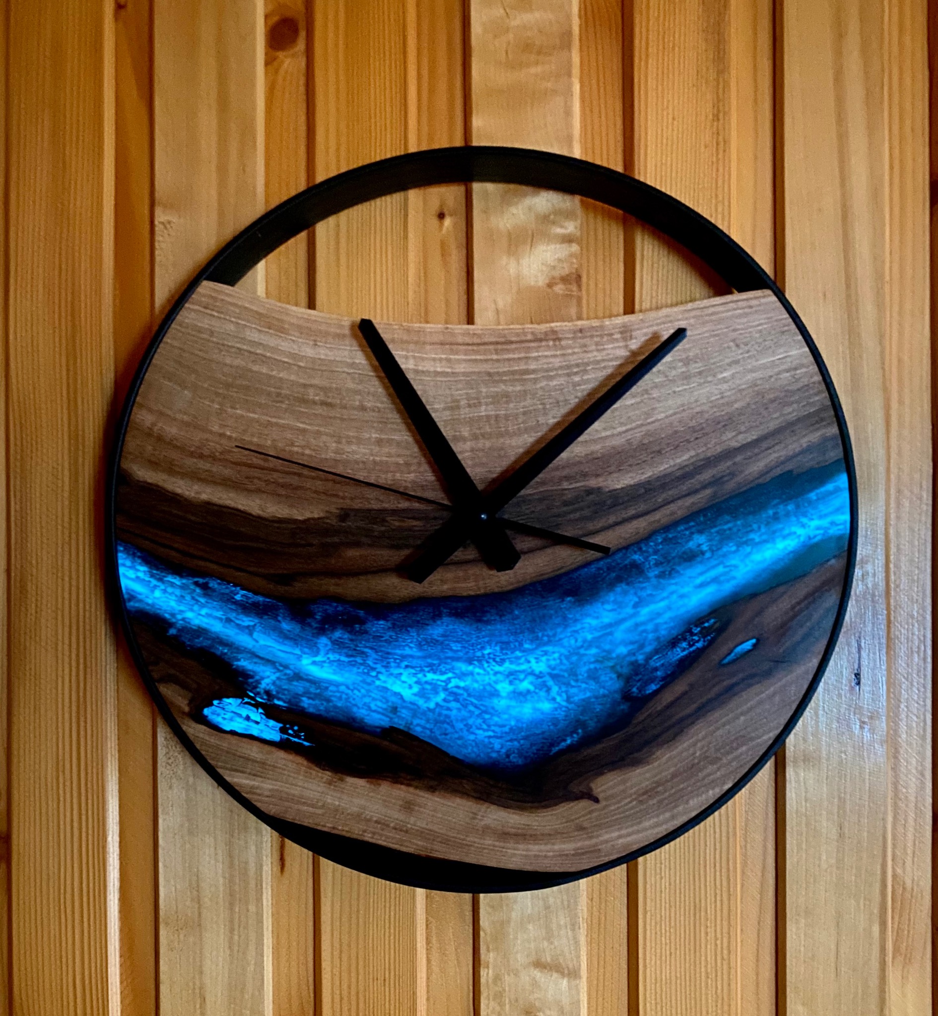 Okrągły zegar z drewna i żywicy epoksydowej, z czarnymi wskazówkami, zawieszony na drewnianej ścianie z paneli. Unikalny design z efektem świecącej rzeki.