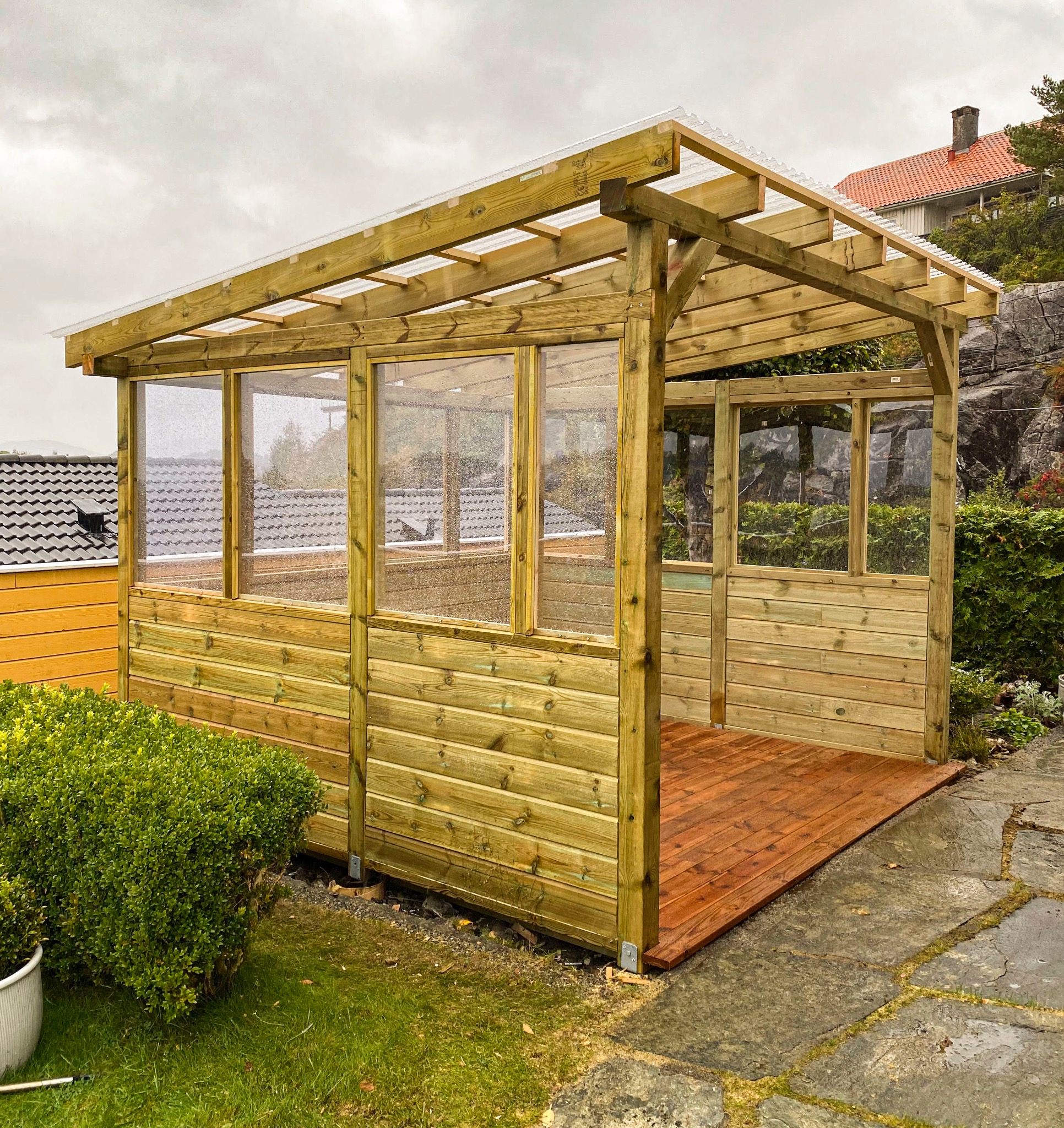 Drewniana pergola z przezroczystym zadaszeniem i osłonami, wykończona na brązowo, na zewnątrz w pochmurny dzień. Widoczne elementy konstrukcyjne i deski podłogowe.