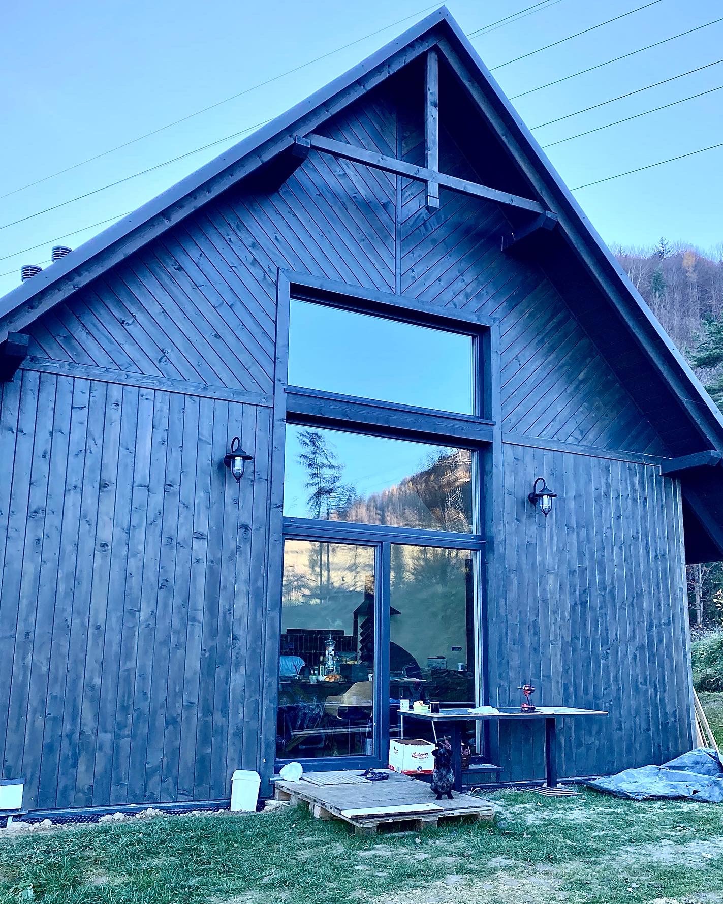 Ciemny, drewniany dom z dużymi oknami i czarnym psem na podeście. Widoczne góry w tle i lampy zewnętrzne. Charakterystyczne pionowe i skośne deski.