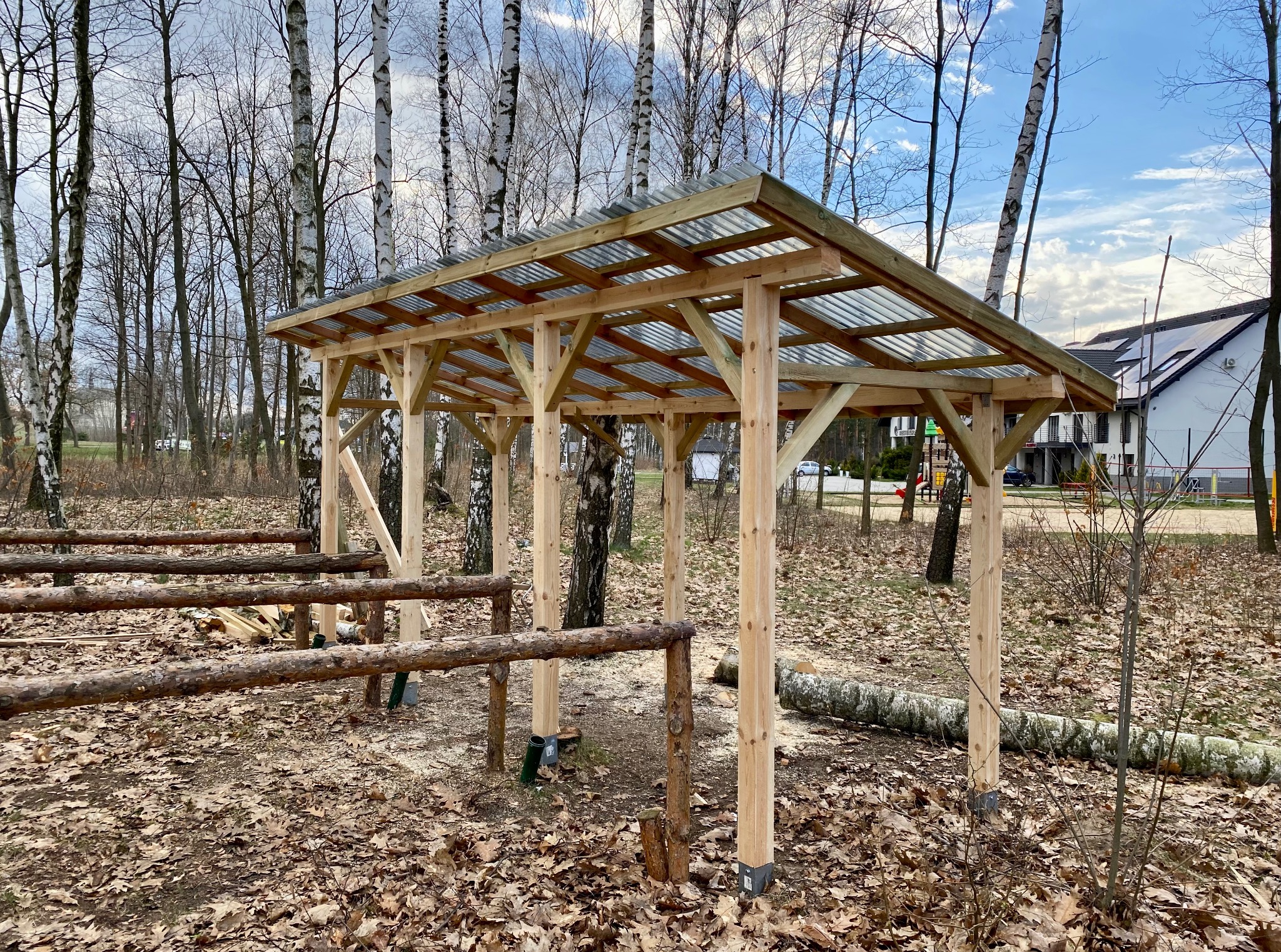 Surowa konstrukcja z drewna, prawdopodobnie altana lub wiata, z przezroczystym zadaszeniem, w otoczeniu drzew i opadłych liści. Widoczne elementy mocujące.