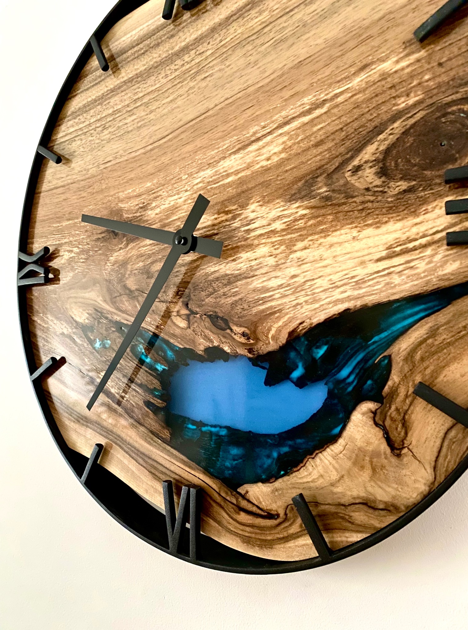 Drewniany zegar z żywicą epoksydową, czarne wskazówki i obramowanie. Unikalny wzór drewna i błękitna żywica tworzą efektowny kontrast. Widoczne detale wykończenia.