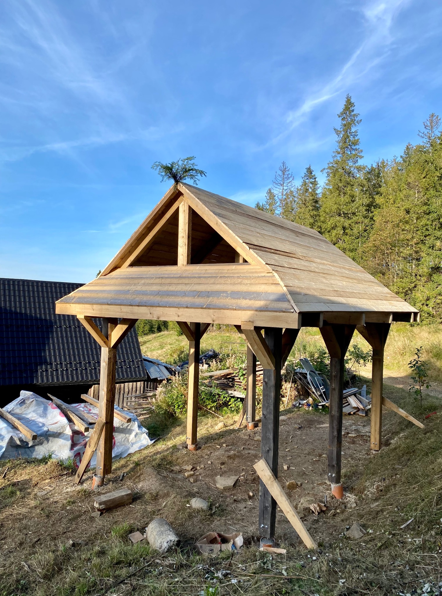 Wznoszona drewniana konstrukcja z dachem w plenerze, widoczne elementy montażowe i narzędzia, w tle las i budynek. Etap budowy z surowego drewna.