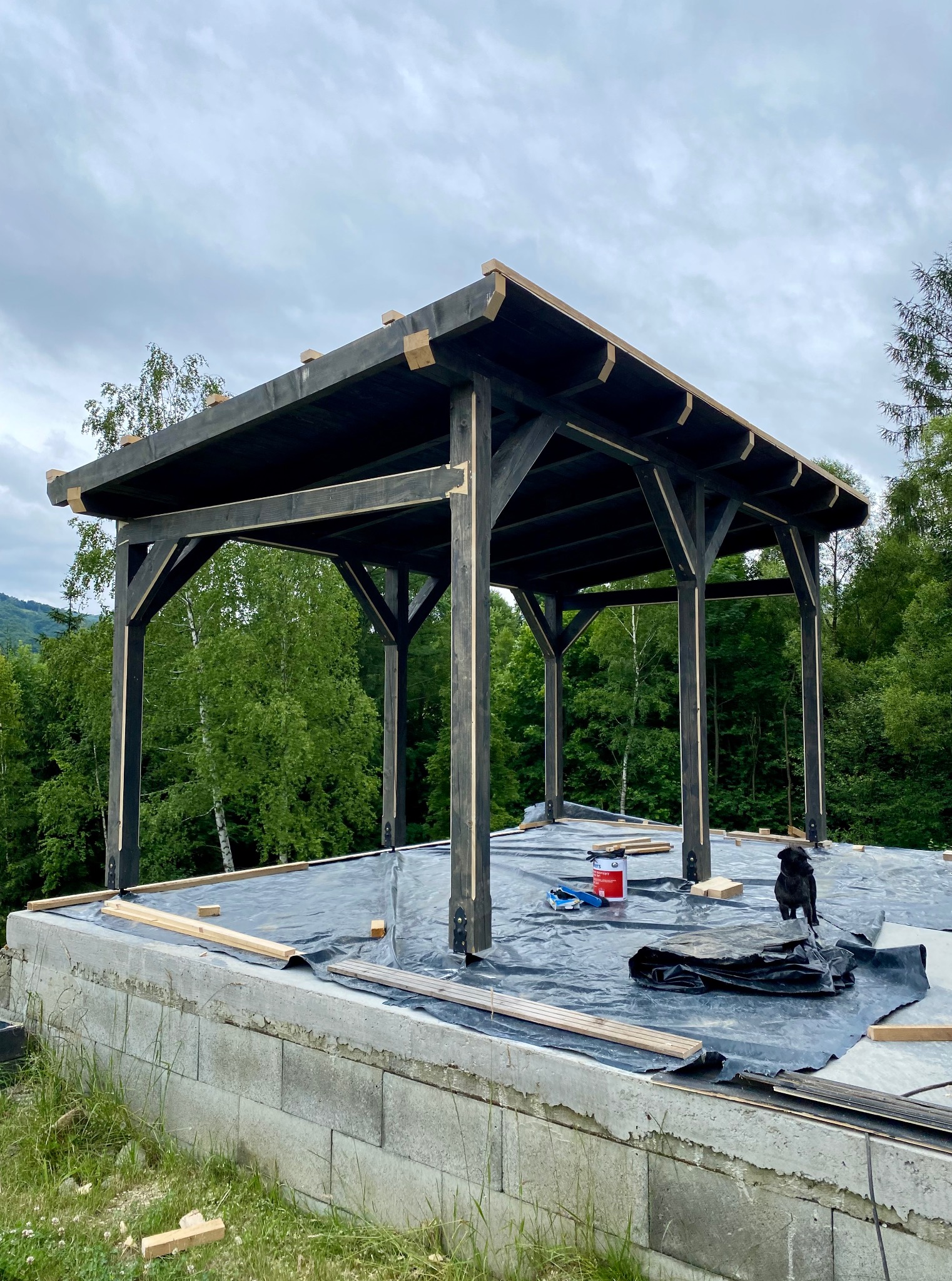 Ciemna, drewniana konstrukcja z widocznymi elementami montażowymi na fundamencie z bloczków, otoczona zielenią. Na dachu leżą narzędzia i puszka farby.