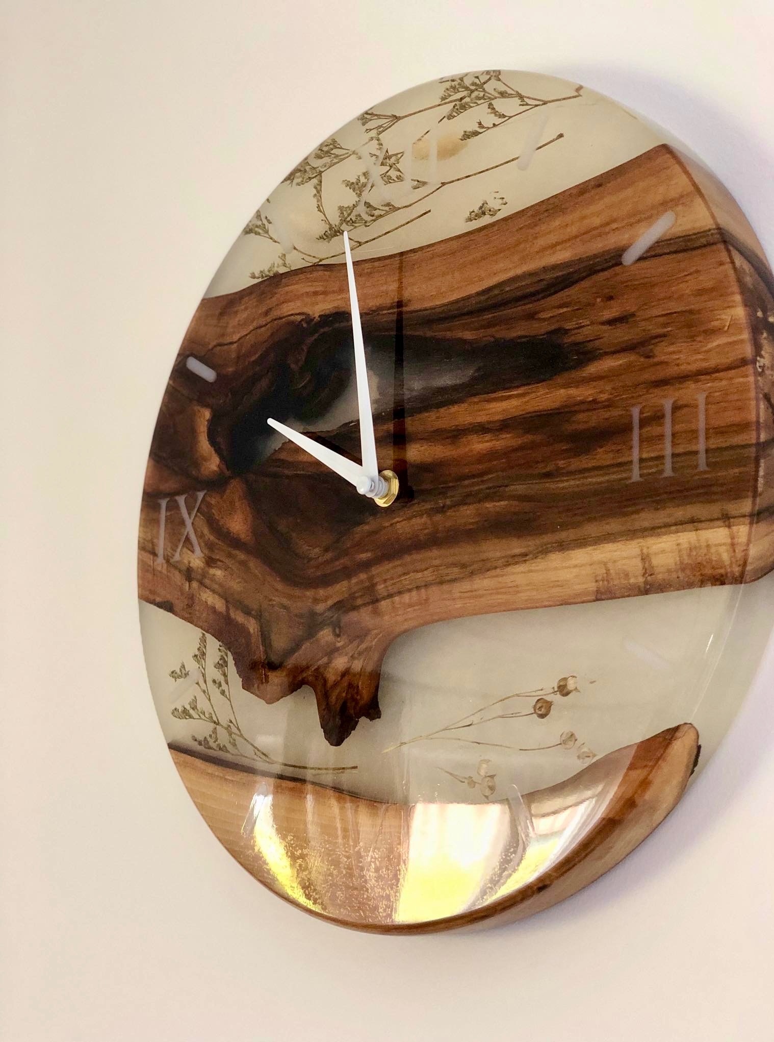 Okrągły zegar ścienny z drewna i żywicy epoksydowej, z zatopionymi suszonymi kwiatami i wyraźnymi oznaczeniami godzin rzymskich i arabskich. Unikatowy design.