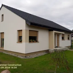 Parterowy dom z ciemną dachówką i jasną elewacją, z widocznym ogrodem i elementami wykończenia zewnętrznego, sfotografowany w pochmurny dzień.