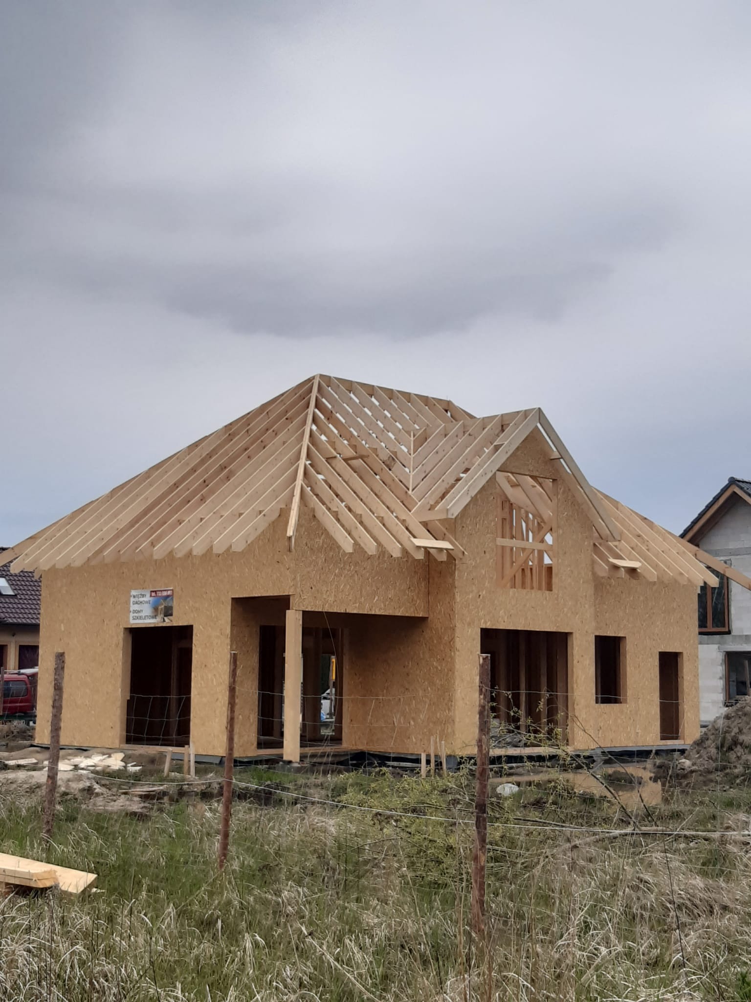 Surowy stan domu szkieletowego z widoczną konstrukcją dachu. Na ścianie budynku naklejka informacyjna. W tle pochmurne niebo i sąsiedni budynek.