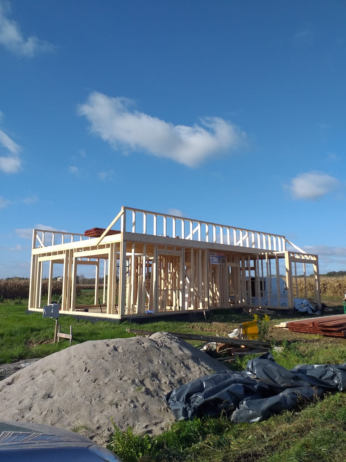 Wznoszenie konstrukcji szkieletowej domu jednorodzinnego na tle błękitnego nieba z chmurami. Widoczny stos piasku na pierwszym planie i elementy konstrukcyjne dachu.