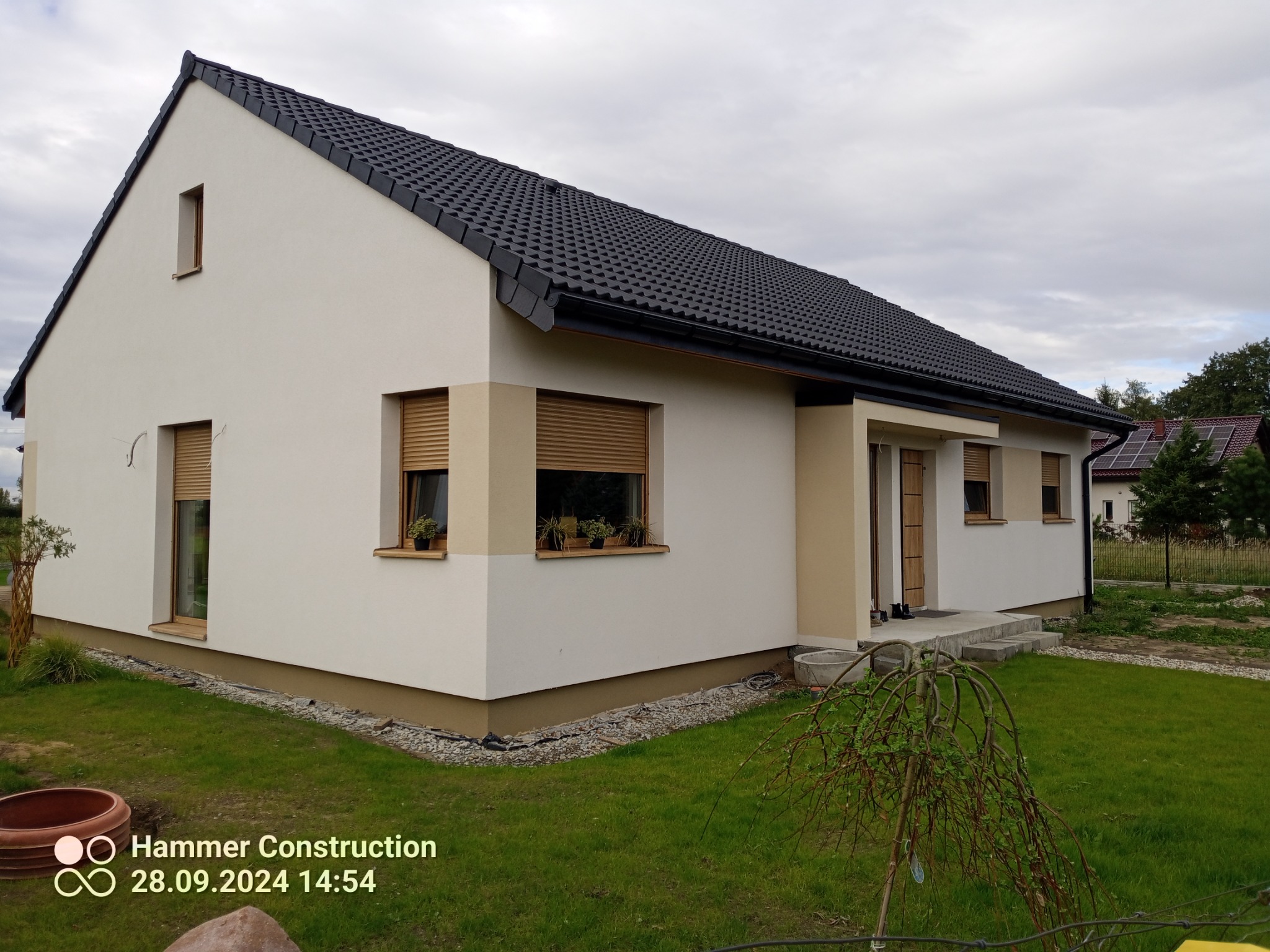 Parterowy dom z ciemną dachówką i jasną elewacją, z widocznym ogrodem i elementami wykończenia zewnętrznego, sfotografowany w pochmurny dzień.