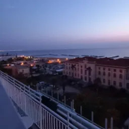 Wieczorny widok z balkonu na miasto nad morzem z budynkami i oświetlonymi ulicami, sfotografowany z góry przy zachodzącym słońcu.