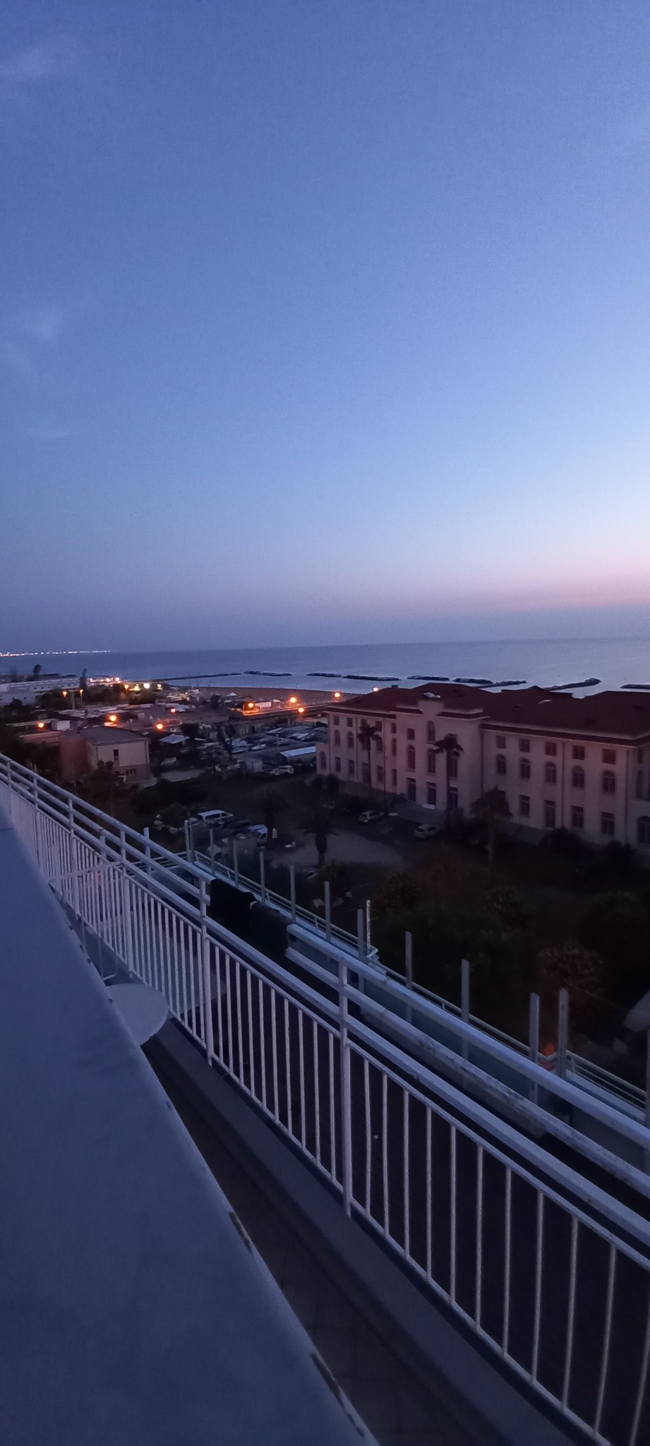 Wieczorny widok z balkonu na miasto nad morzem z budynkami i oświetlonymi ulicami, sfotografowany z góry przy zachodzącym słońcu.