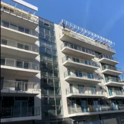 Wielopiętrowy budynek w trakcie prac elewacyjnych. Widoczne rusztowania, balkony z szklanymi balustradami i fragmenty ocieplenia. Częściowo zakończone prace.