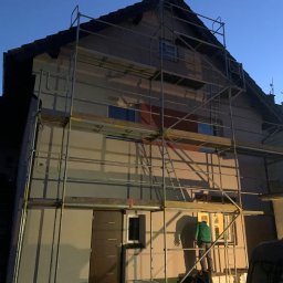 Yaroslav Dudych-Bondarenko D.B. Elewacja Expert - Dom w rusztowaniach podczas prac elewacyjnych o zmierzchu. Widoczny pracownik w zielonej bluzie na rusztowaniu. Częściowo otynkowana ściana.