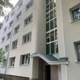 Odnowiona elewacja bloku mieszkalnego z jasną fasadą, białymi oknami i przeszkloną klatką schodową, sfotografowana z dołu w słoneczny dzień.