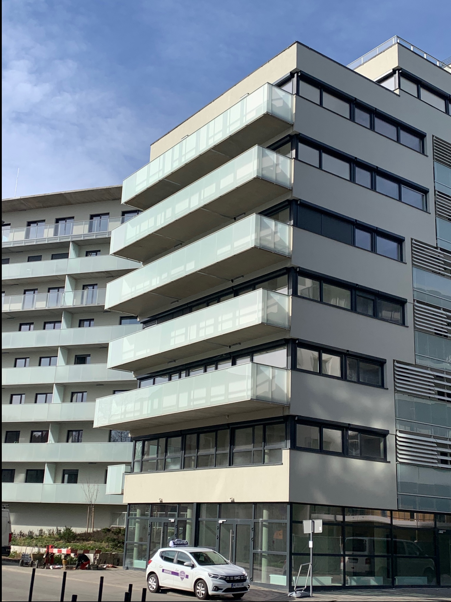 Nowoczesny budynek wielorodzinny z balkonami o szklanych balustradach, widoczny z perspektywy ulicy z zaparkowanym samochodem. Budynek w jasnej kolorystyce.