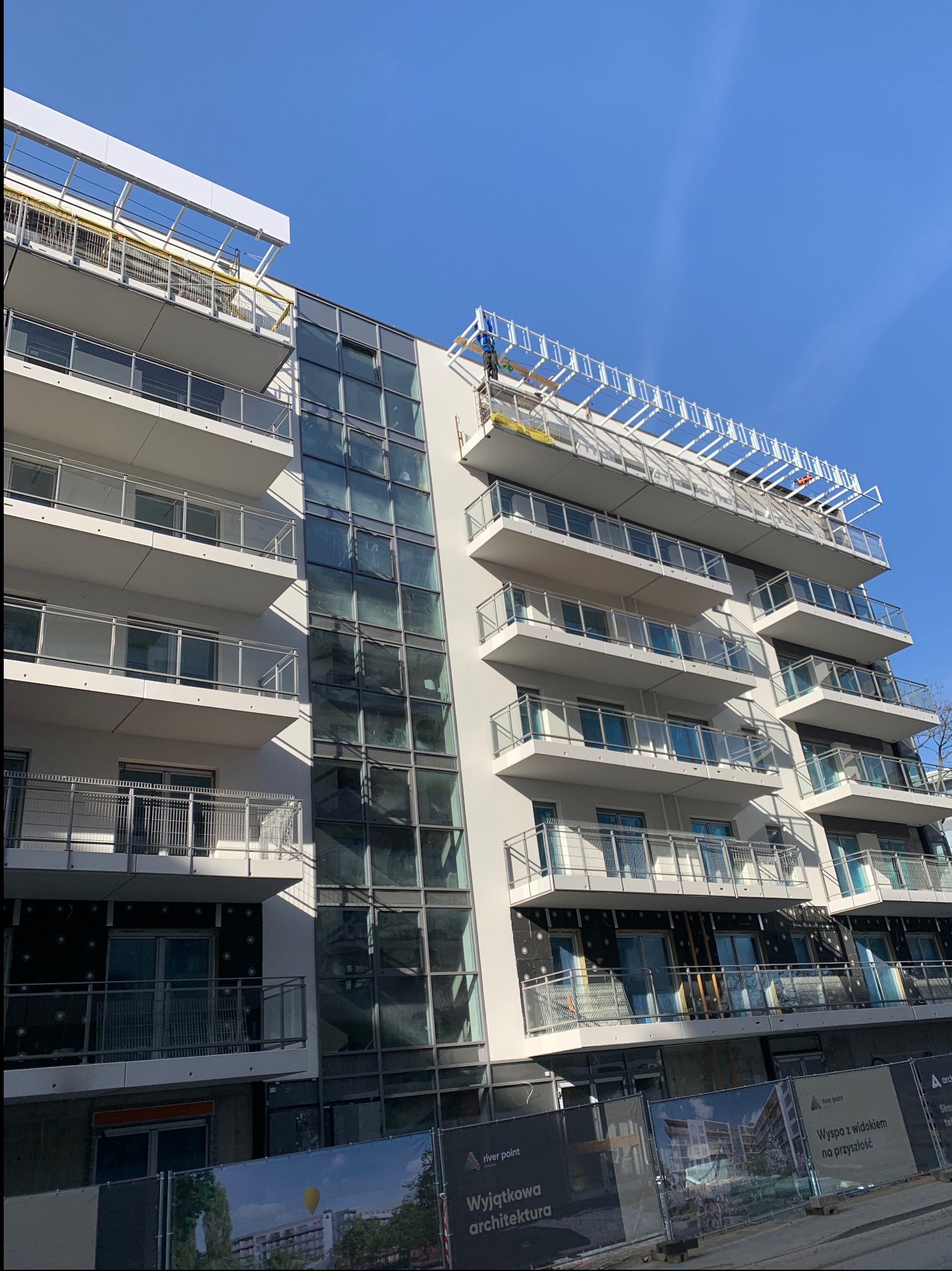 Wielopiętrowy budynek w trakcie prac elewacyjnych. Widoczne rusztowania, balkony z szklanymi balustradami i fragmenty ocieplenia. Częściowo zakończone prace.