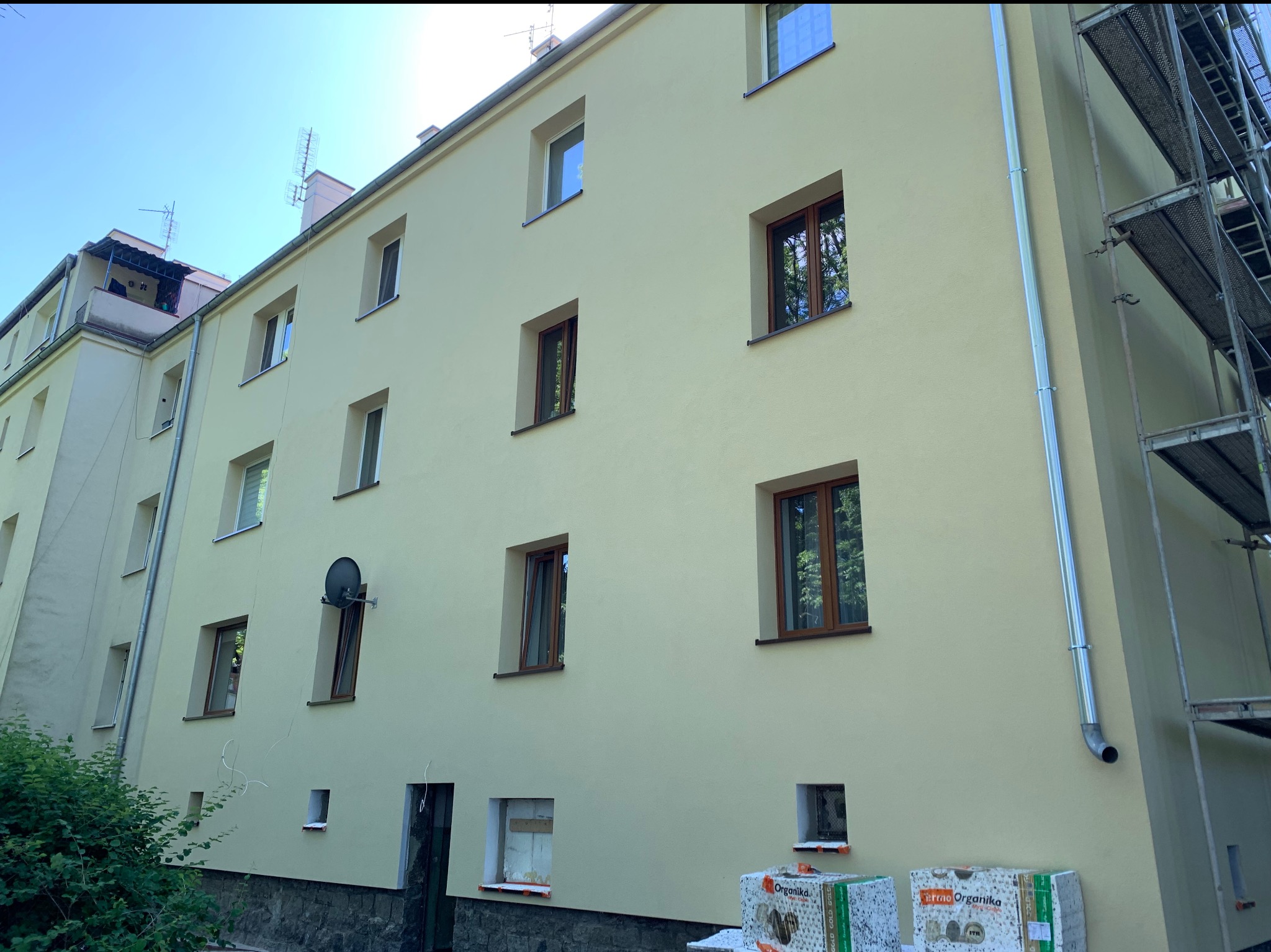 Elewacja budynku po tynkowaniu, z widocznymi oknami i rusztowaniem po prawej stronie. Pakiety styropianu leżą u dołu ściany. Jasne kolory.