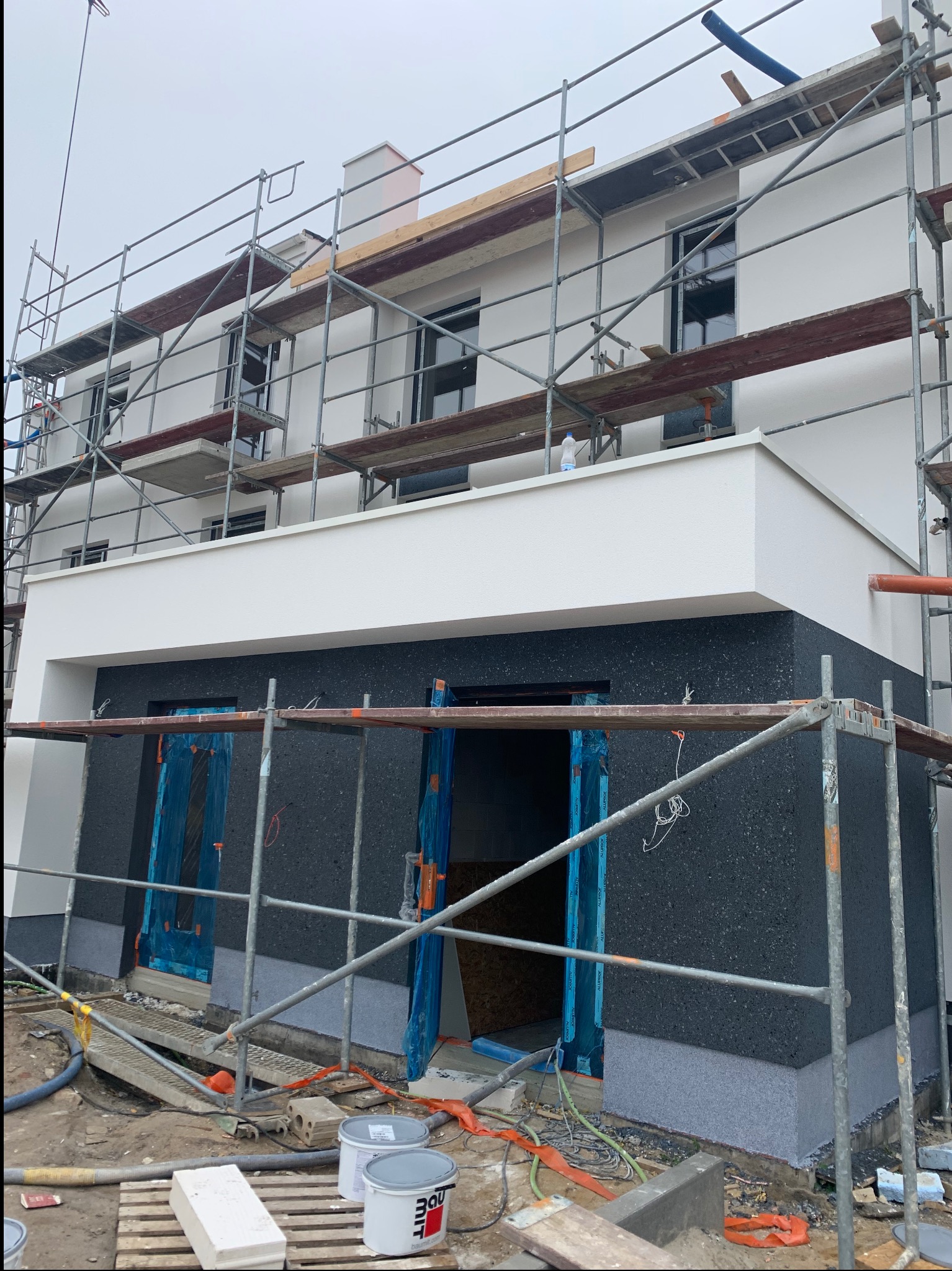 Dwukondygnacyjny dom w trakcie prac elewacyjnych: rusztowania, częściowo otynkowana ściana w ciemnym kolorze, zabezpieczone okna i drzwi folią.