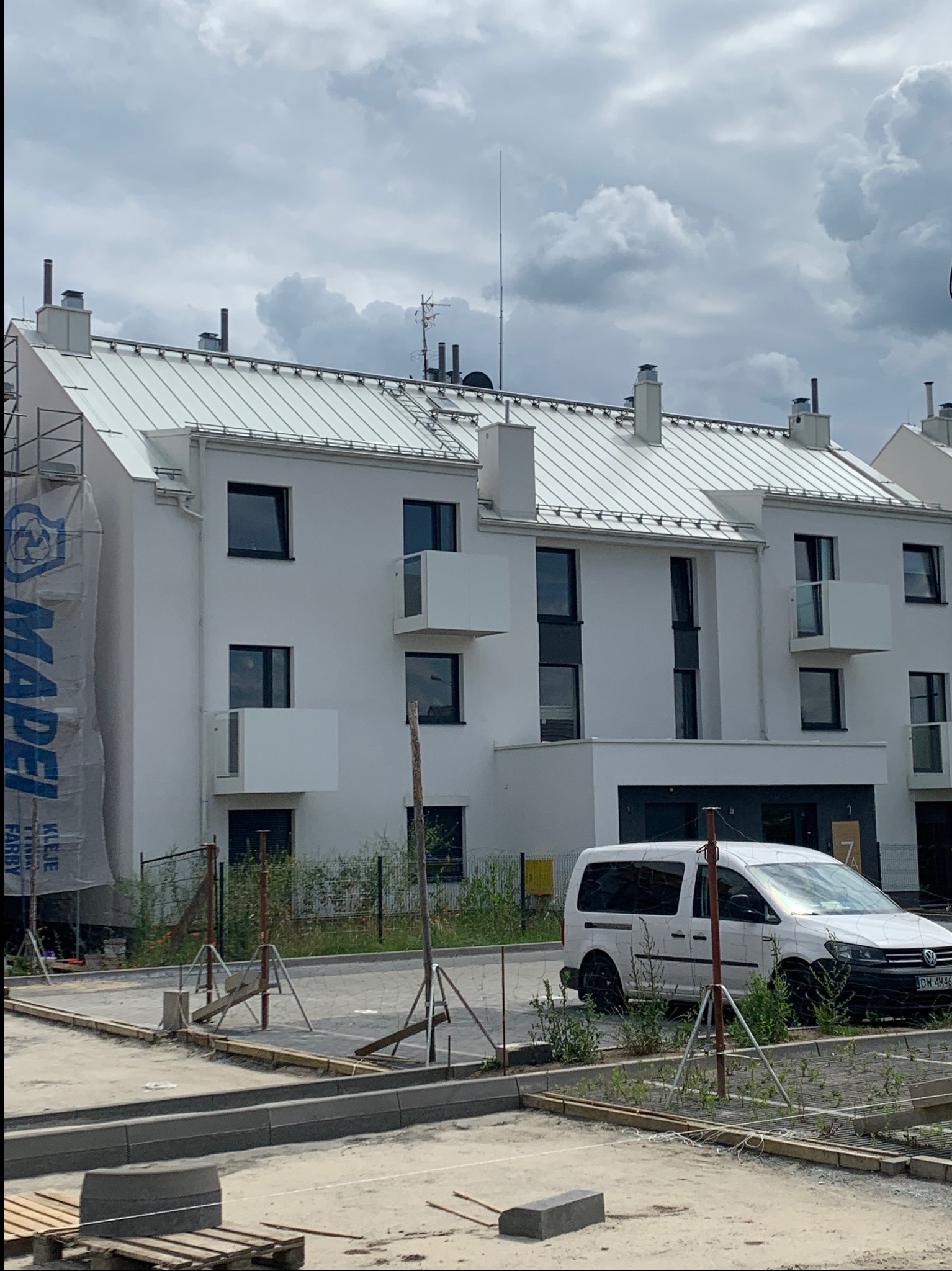 Nowoczesny, trzypiętrowy budynek z białymi elewacjami i metalowym dachem, częściowo zasłonięty banerem reklamowym. Na pierwszym planie widoczne prace brukarskie.