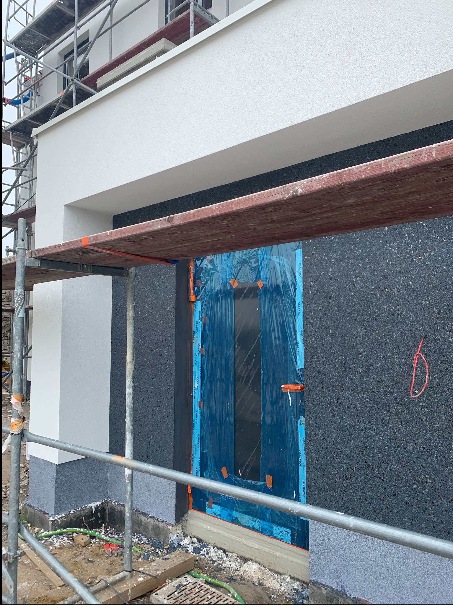 Budynek w trakcie ocieplania: czarny styropian na elewacji, okno zabezpieczone folią, rusztowanie. Fragment budynku z widocznym ociepleniem i przygotowaniem do tynkowania.