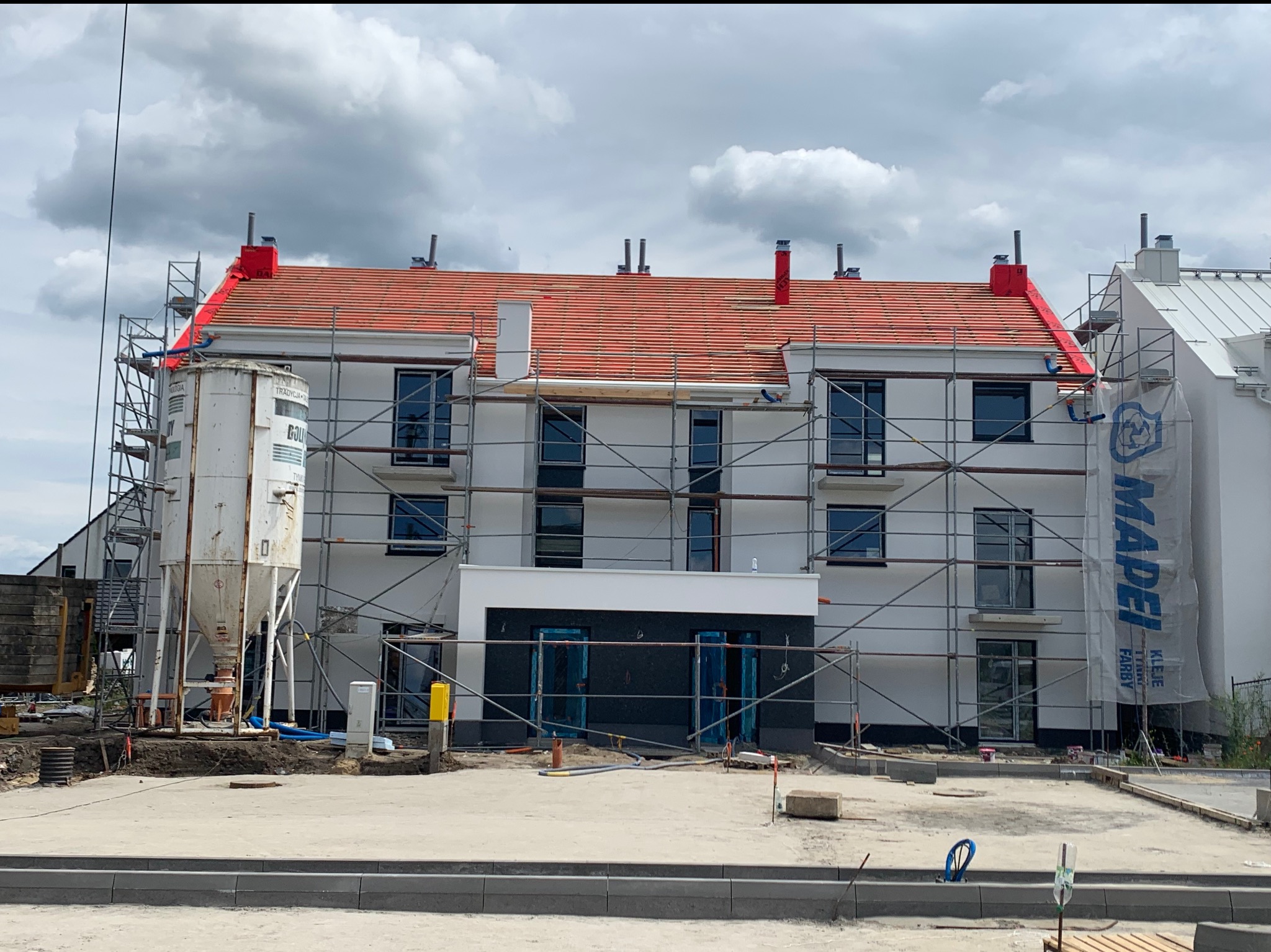 Wielopiętrowy budynek w trakcie prac elewacyjnych, otoczony rusztowaniami i sprzętem budowlanym. Widoczny zbiornik na zaprawę i częściowo zakryta elewacja sąsiedniego budynku.