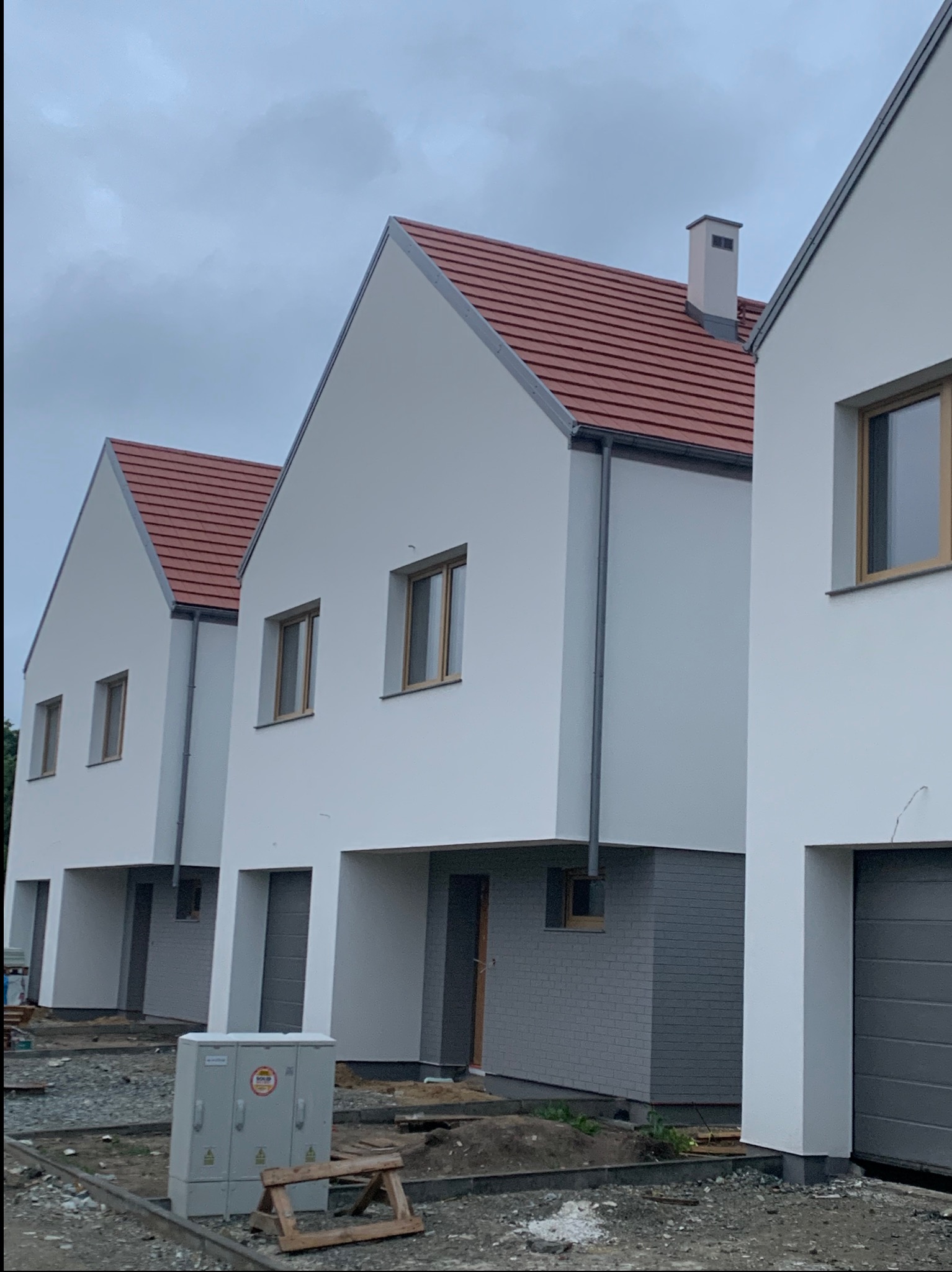 Trzy identyczne, nowo wybudowane domy z białymi elewacjami i czerwoną dachówką, widziane z perspektywy ulicy w pochmurny dzień. W tle szare niebo.