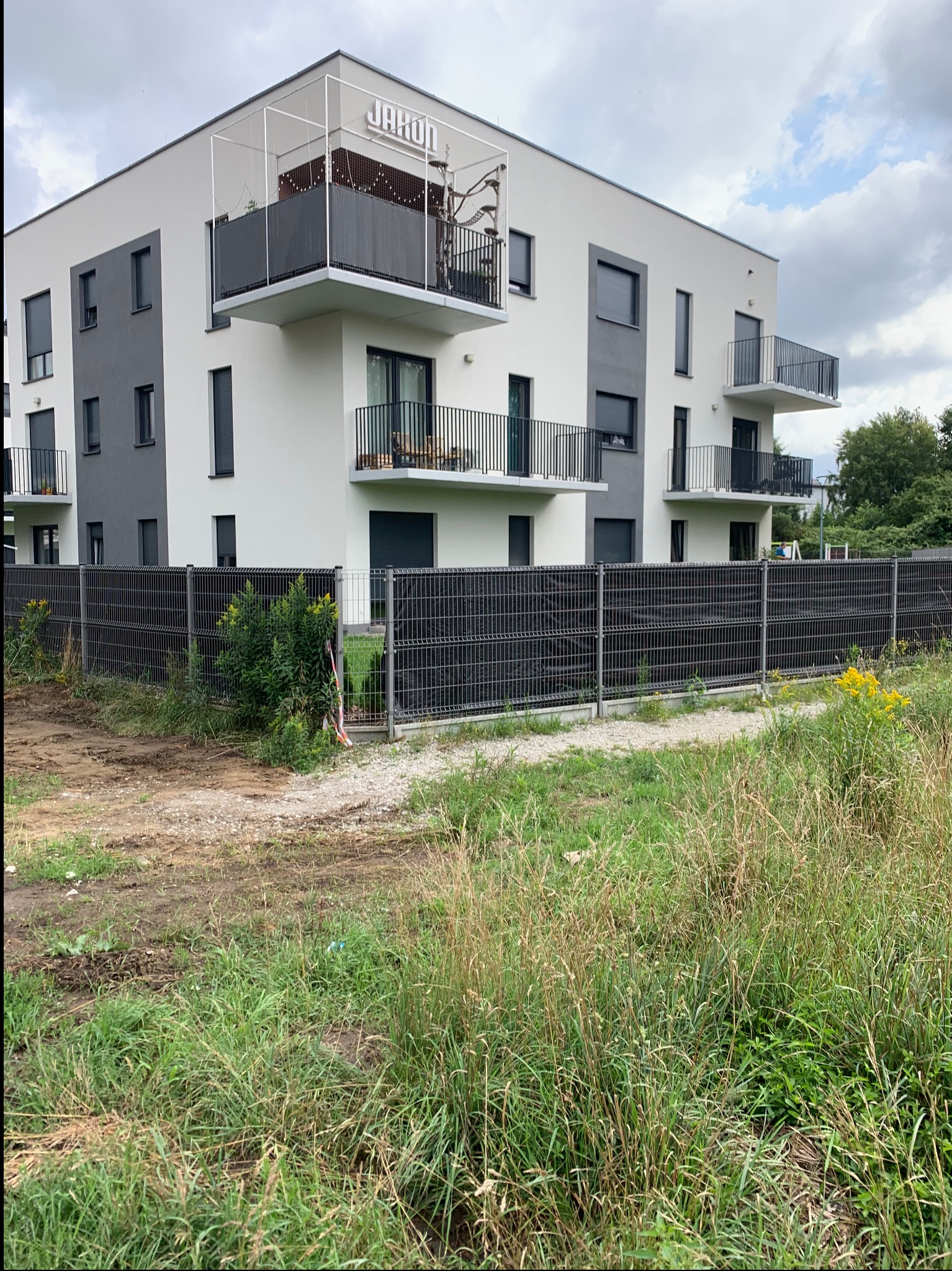 Nowoczesny budynek wielorodzinny z białoszarą elewacją i balkonami z czarnymi balustradami, ogrodzony metalową siatką, z widoczną zielenią w tle i na pierwszym planie.