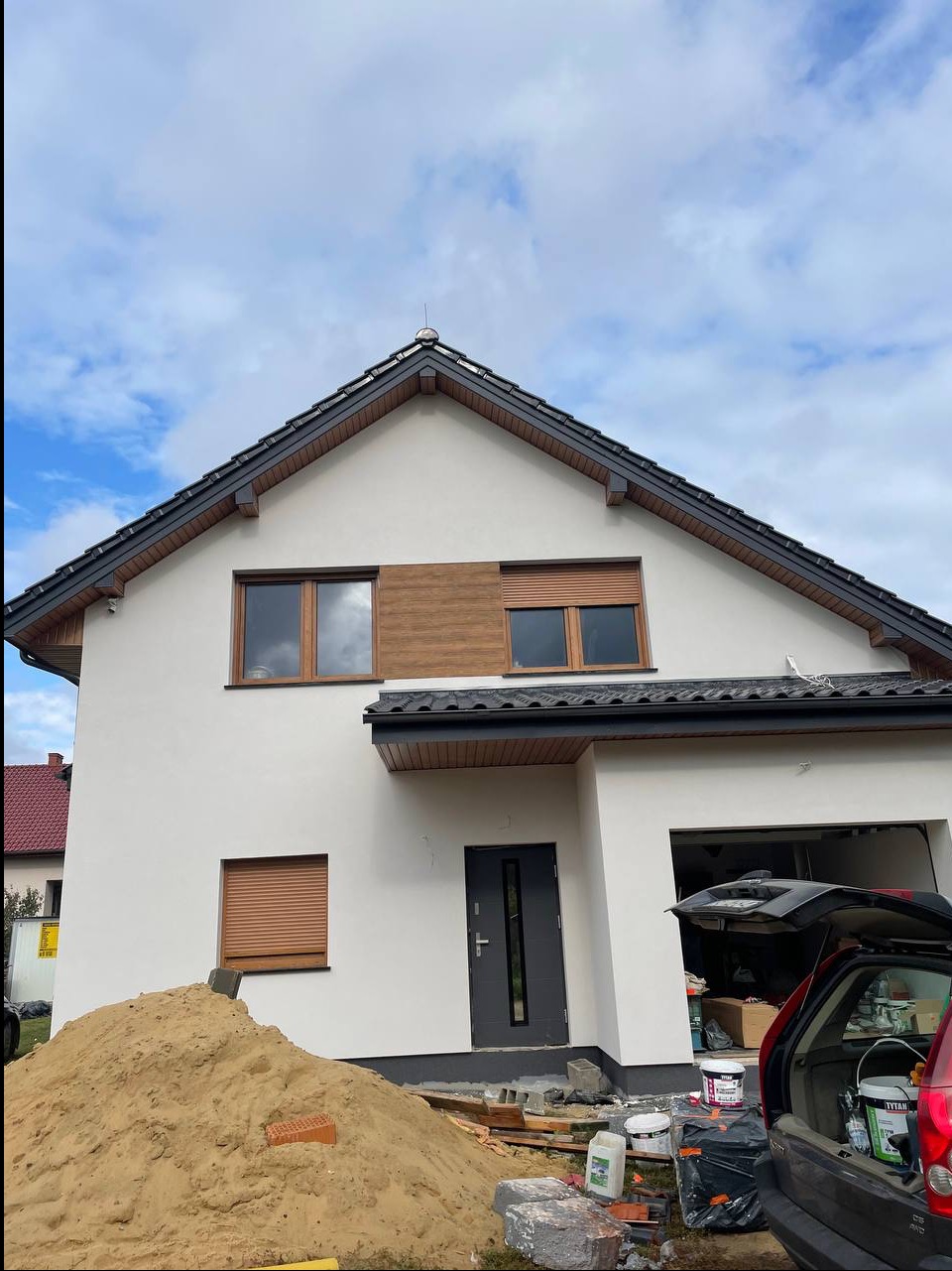 Dwukondygnacyjny dom z jasną elewacją, drewnianymi akcentami i roletami. W tle błękitne niebo i sterta piasku przed budynkiem.