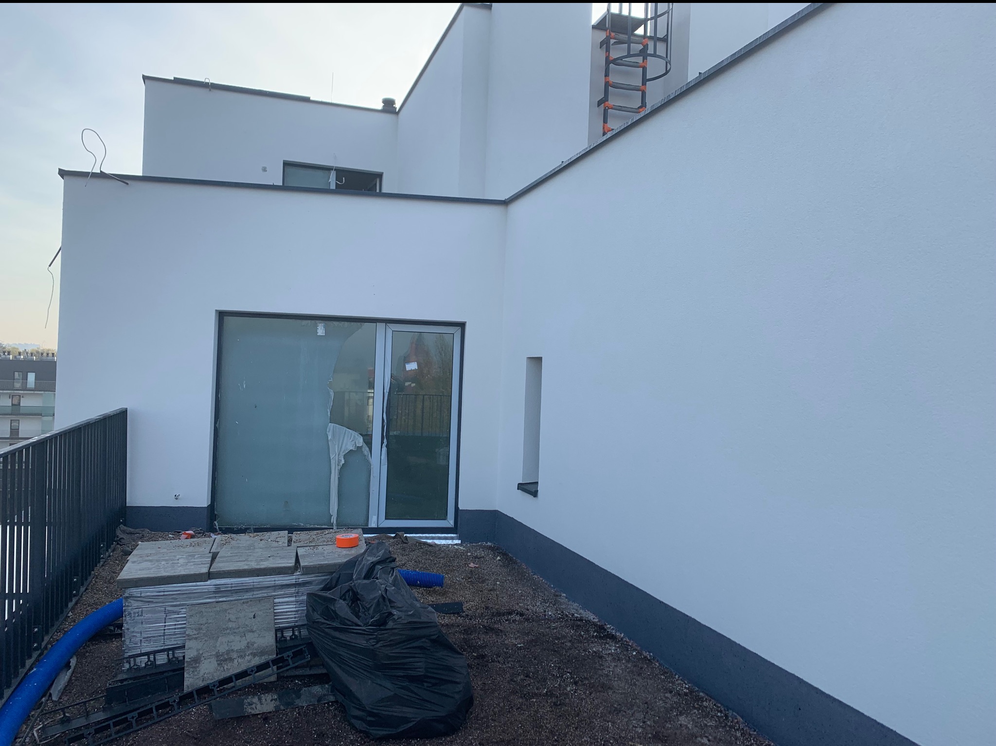 Biała elewacja budynku z wyjściem na taras. Widoczne materiały budowlane i drabina na dachu. Świeżo otynkowana powierzchnia.