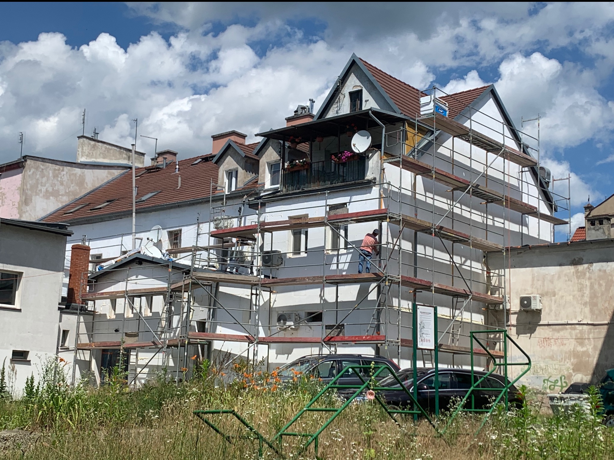 Ocieplanie budynku mieszkalnego w trakcie prac, widoczne rusztowania, pracownik i elementy ocieplenia. W tle błękitne niebo z chmurami.