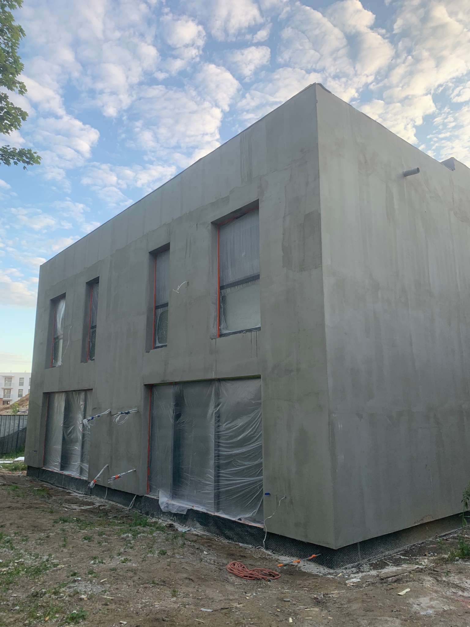 Surowy budynek z szarą elewacją w trakcie tynkowania. Okna zabezpieczone folią. Widoczne przewody na ścianie. Błękitne niebo z chmurami w tle.